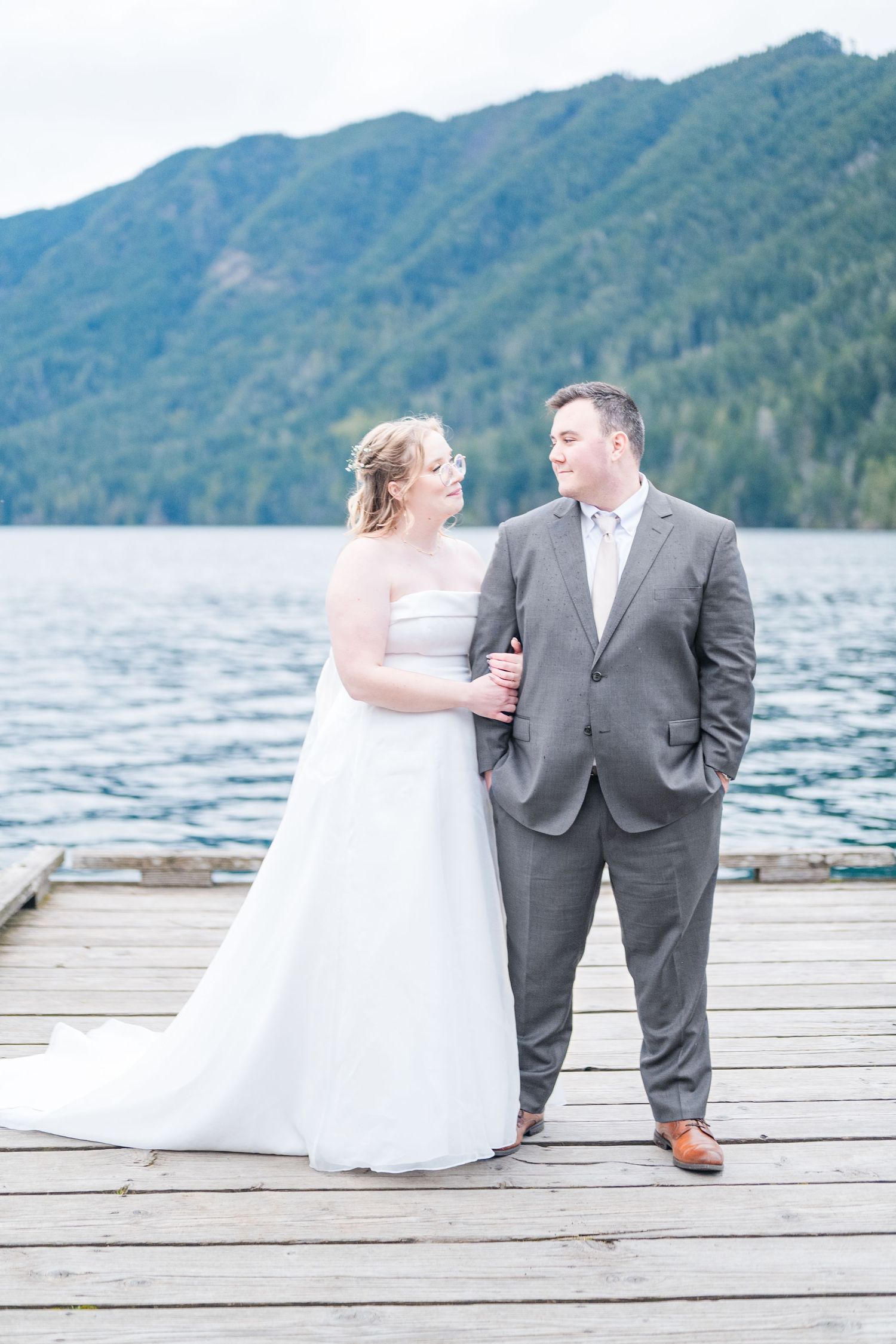 Bride and groom pose on a wooden dock with a serene mountain lake backdrop on their wedding day.
