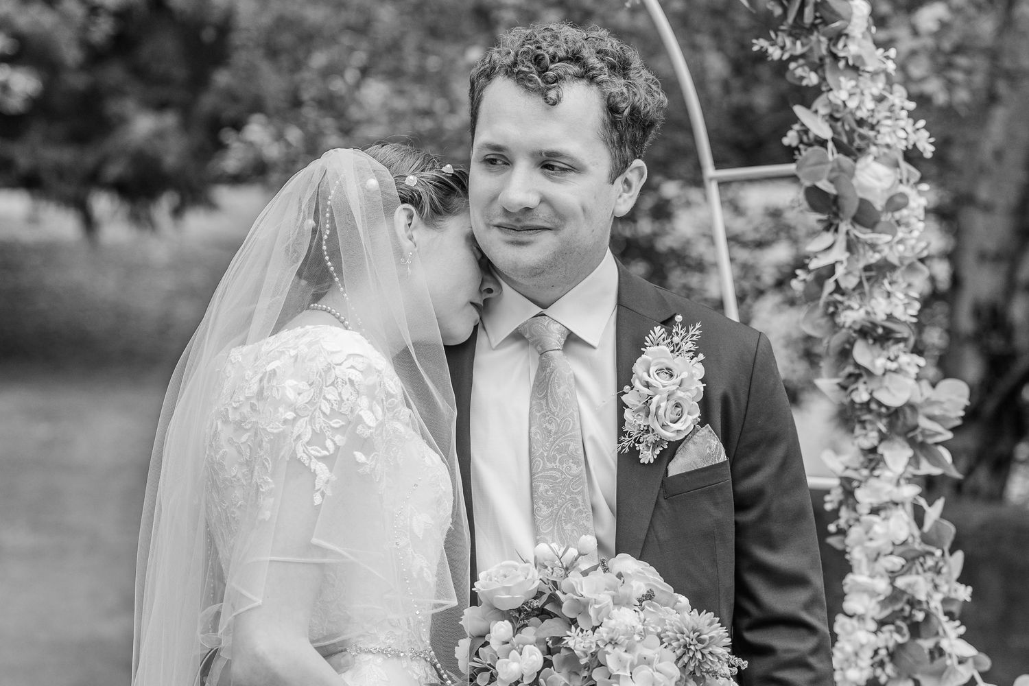 Black and white wedding photo of bride leaning on groom near floral arch, bride holding bouquet in lace dress with veil.
