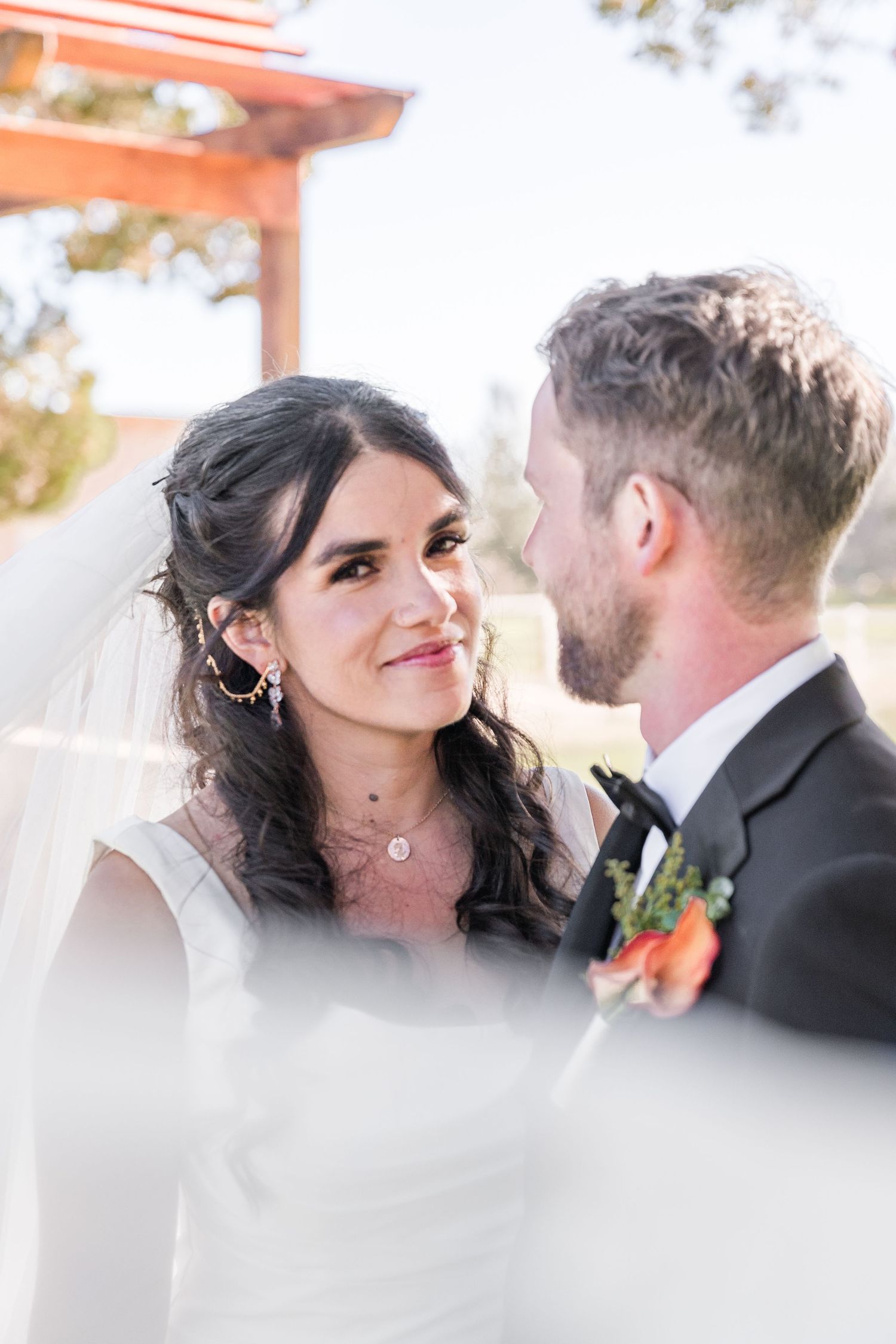 Bride in white veil smiles lovingly at groom in black tuxedo with orange boutonniere during outdoor wedding ceremony.