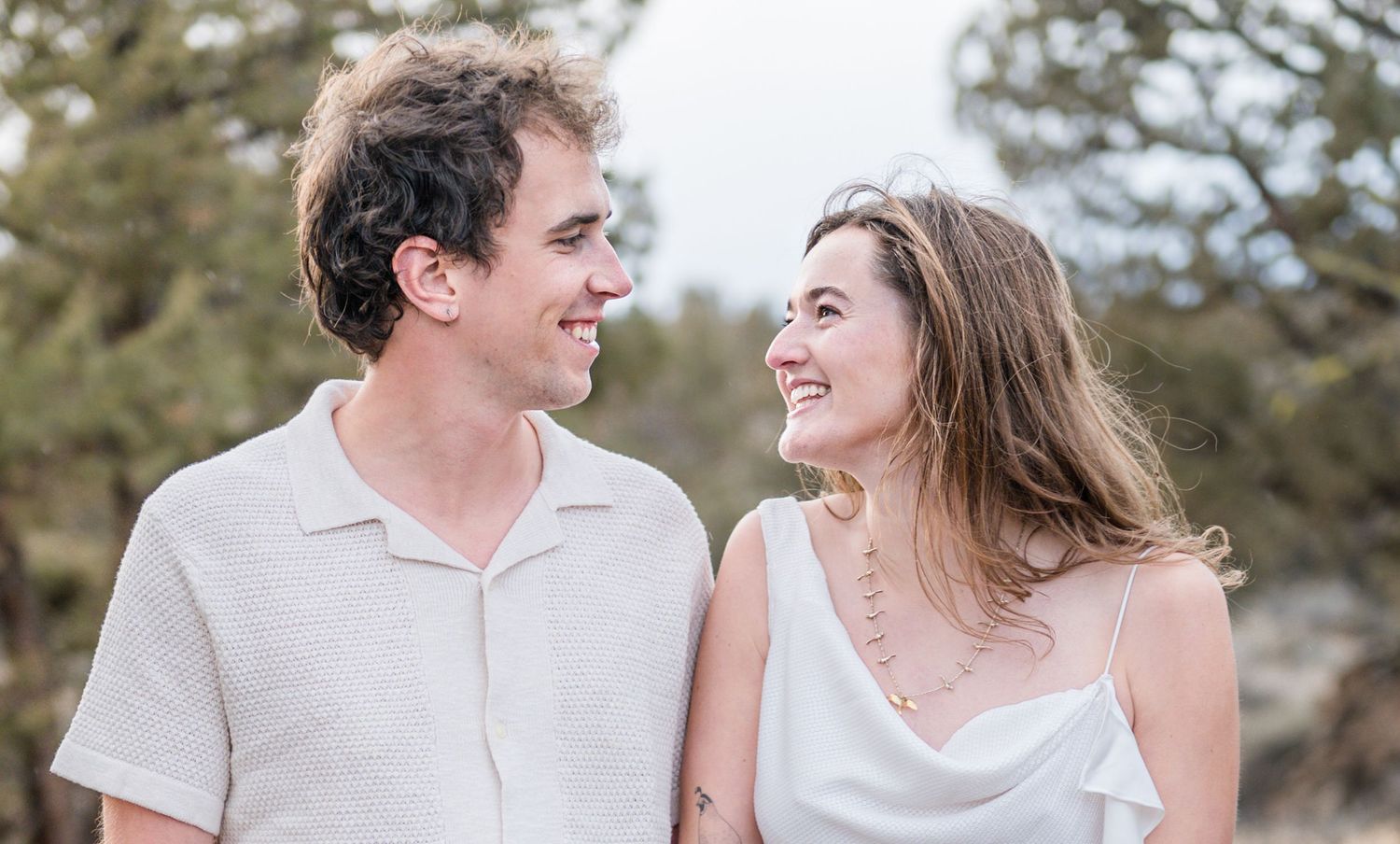 A smiling couple in white outfits sharing a joyful moment outdoors with pine trees in the background.