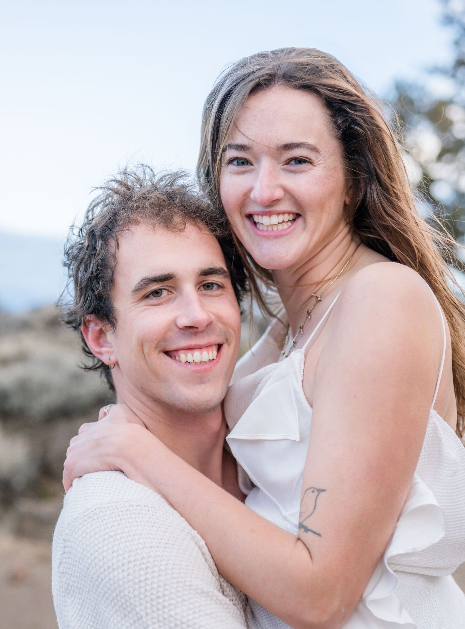 A smiling couple in white outfits embracing outdoors with a scenic natural background.