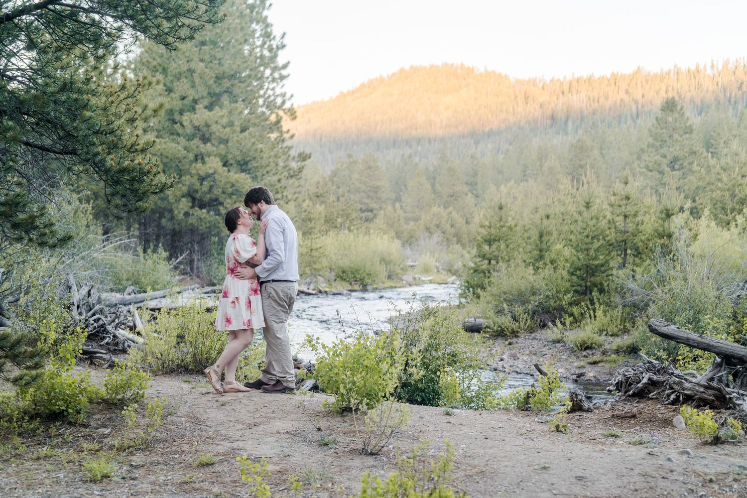 Couple sharing a romantic kiss near a mountain stream surrounded by pine trees at sunset.