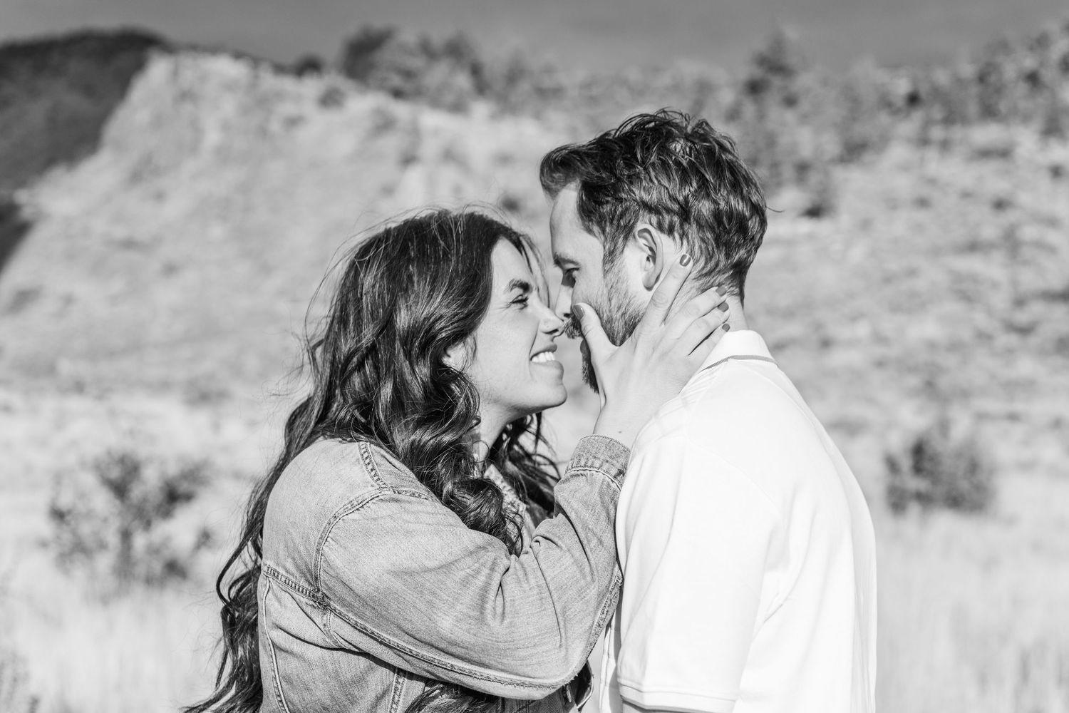 Black and white photo of a romantic couple sharing an intimate moment outdoors with scenic hills in the background.