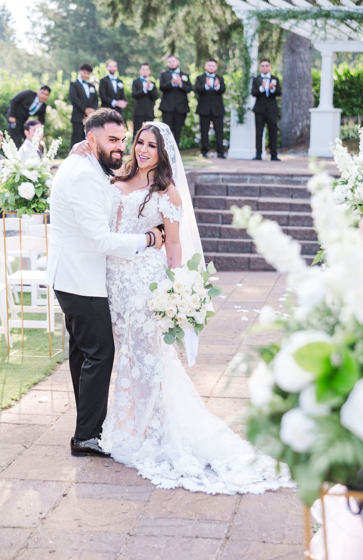 Bride and groom share a romantic moment at outdoor wedding ceremony with white floral arrangements and groomsmen in background.