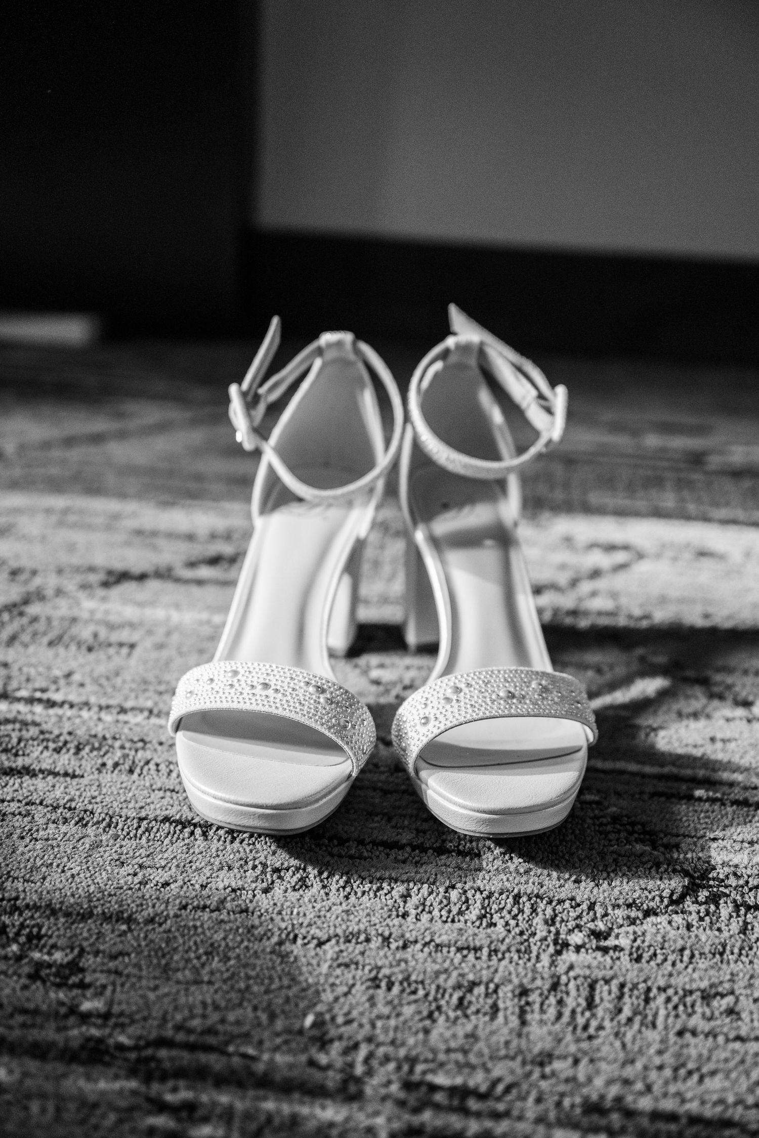 Elegant white bridal heels with rhinestone embellishments and ankle straps displayed on a textured carpet in black and white.
