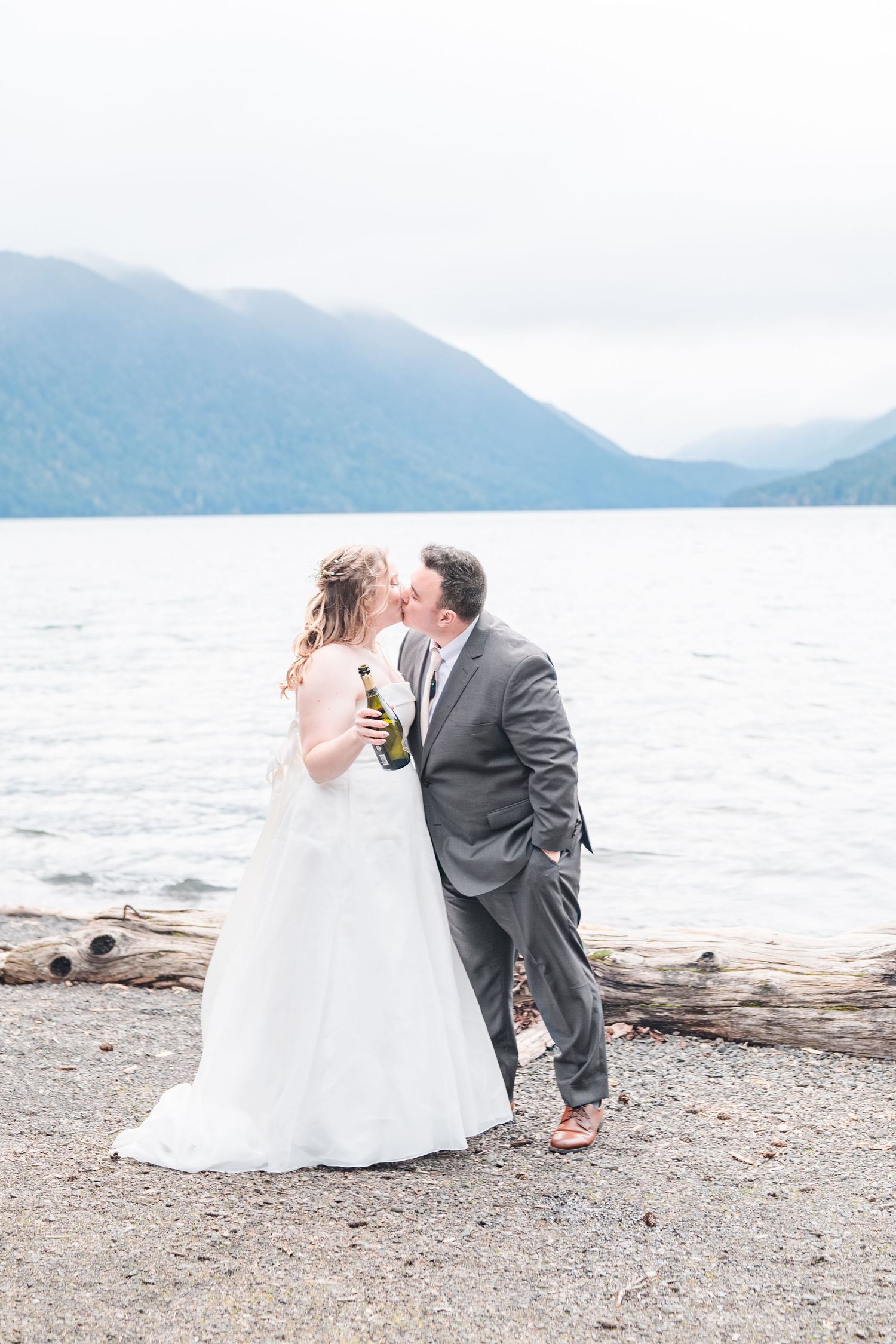 Bride in a white gown and groom in a gray suit kiss on a lakeside beach holding a champagne bottle, with misty mountains behind them.