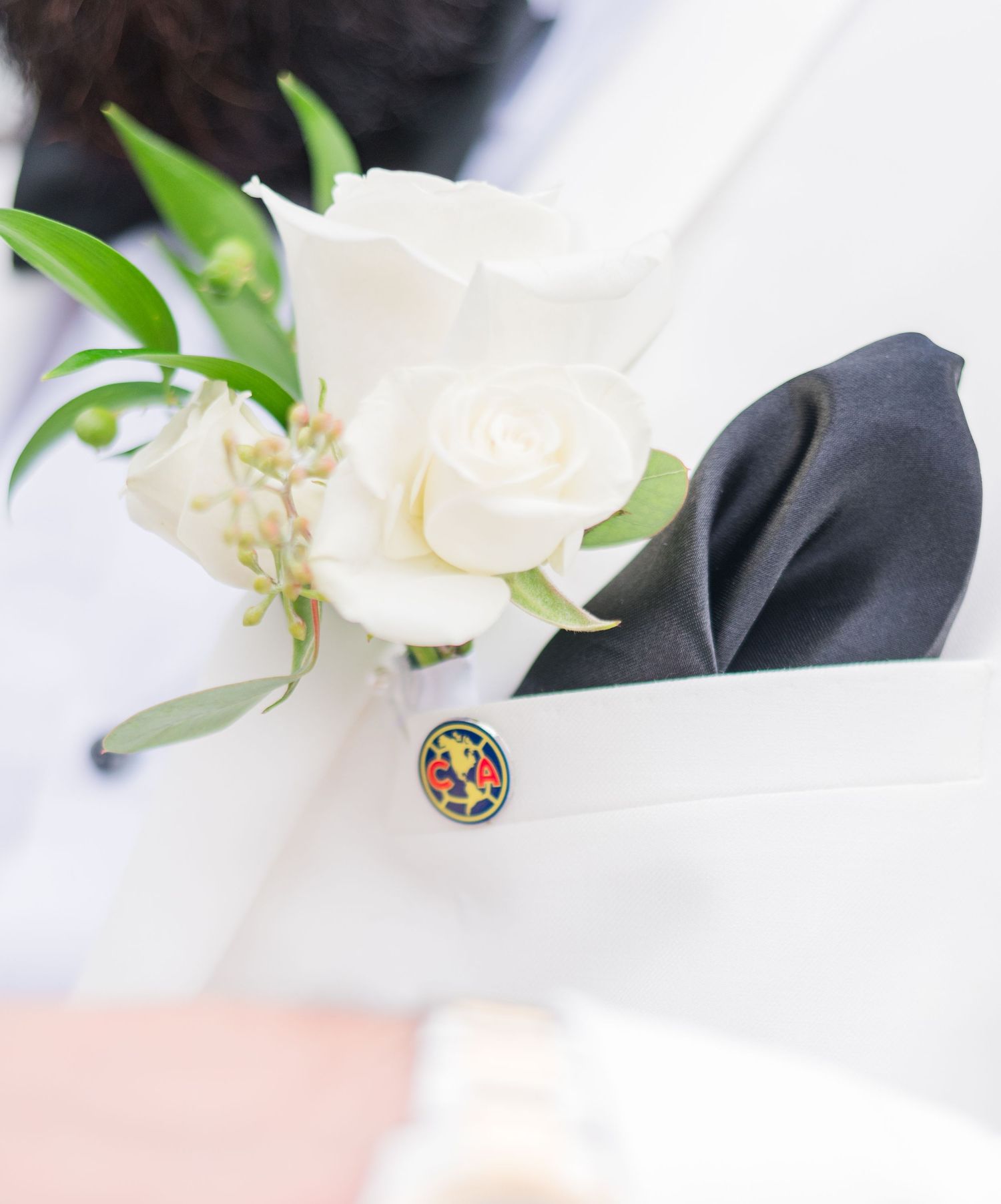 White rose boutonniere with greenery on a white lapel, accented by a dark pocket square and decorative pin.