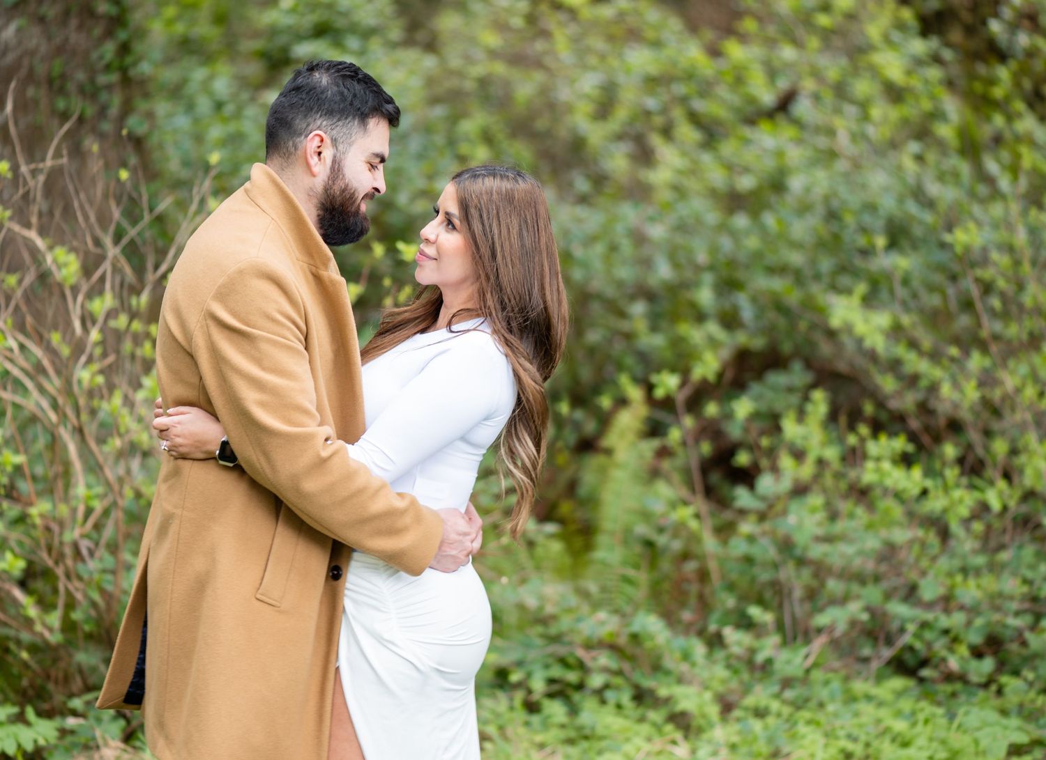 Couple embracing outdoors, woman in white maternity dress and man in tan coat, surrounded by lush green foliage.