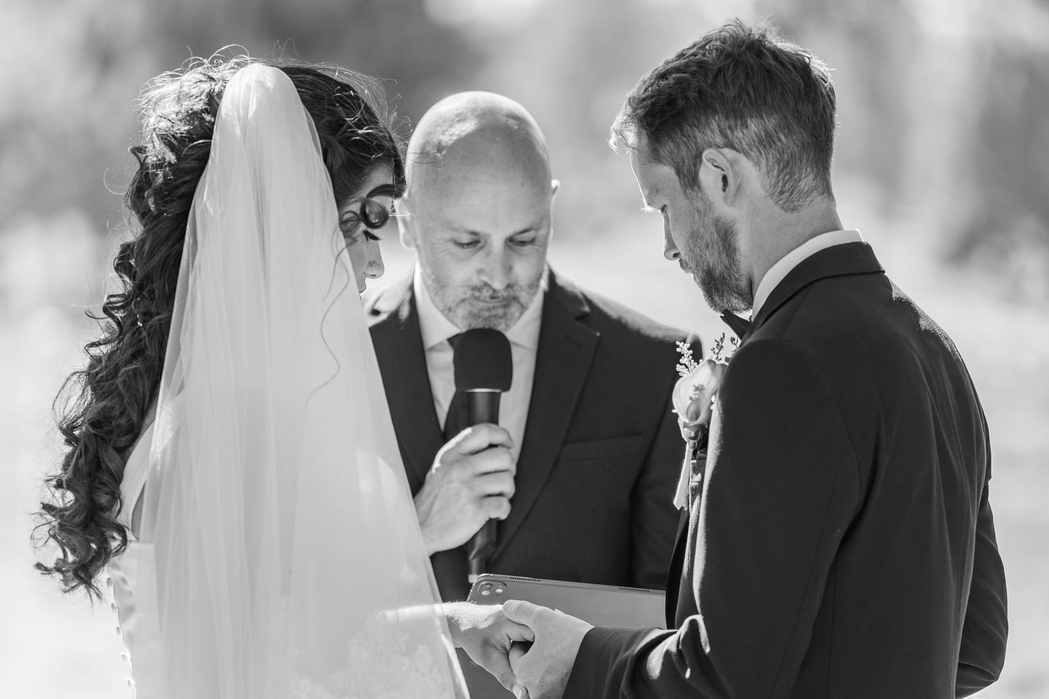 Black and white wedding ceremony photo of bride and groom exchanging vows with officiant holding microphone.