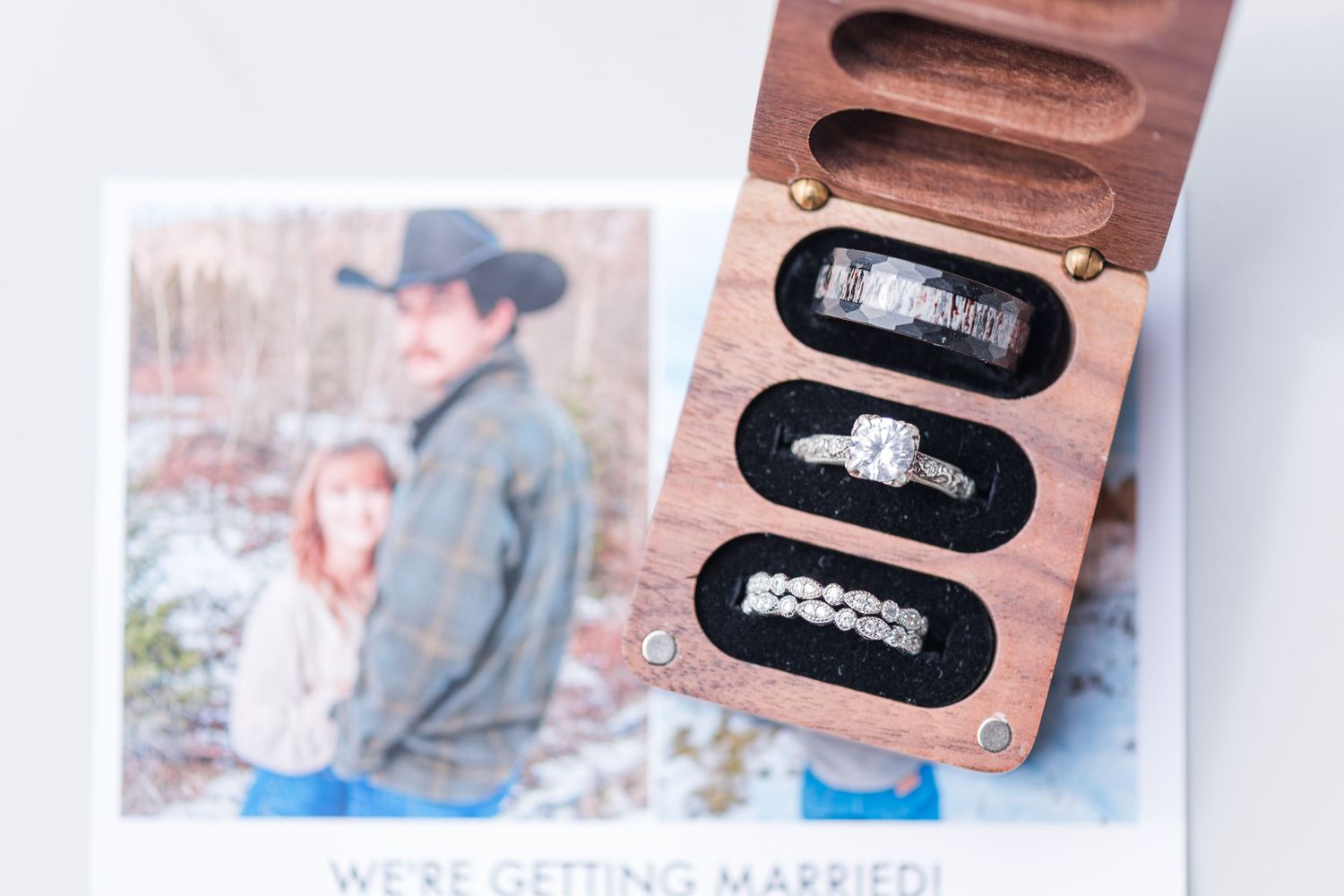 Wooden ring box with wedding bands beside a We're Getting Married card featuring an engaged couple in a snowy outdoor setting.