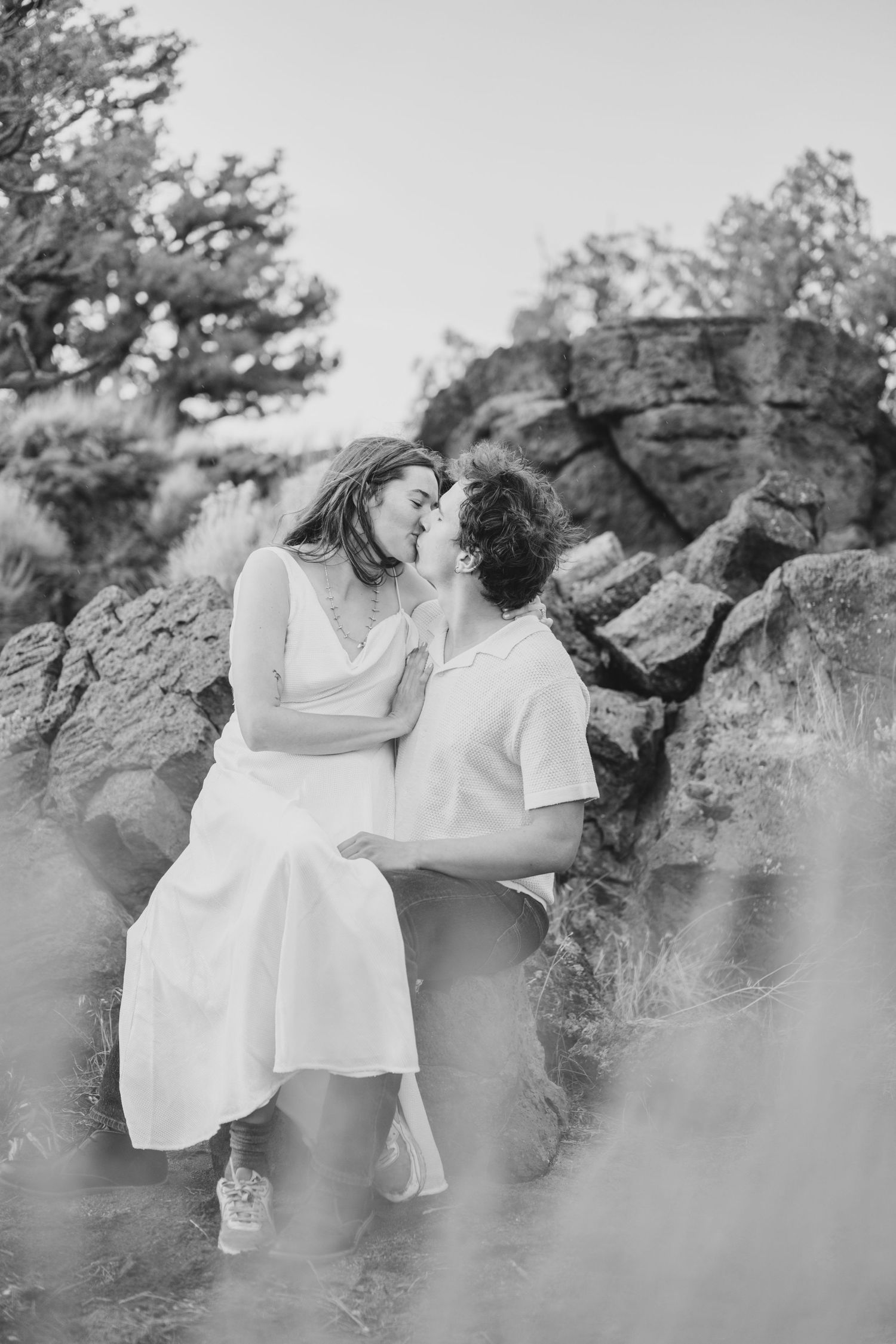 Black and white photo of a couple sharing a romantic kiss on rocky terrain surrounded by trees and nature.