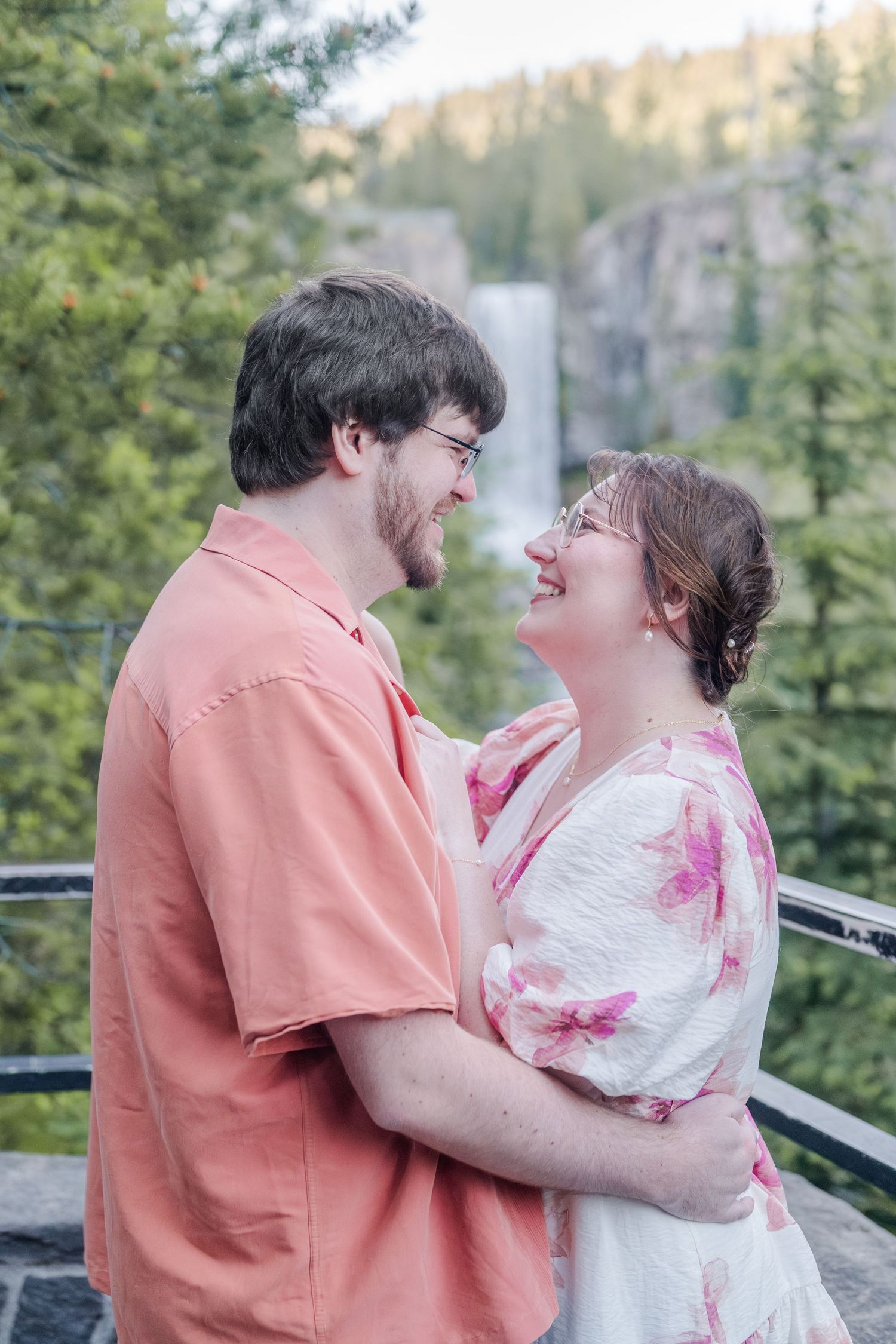A couple embraces and smiles at each other on a scenic overlook with a waterfall and trees in the background.