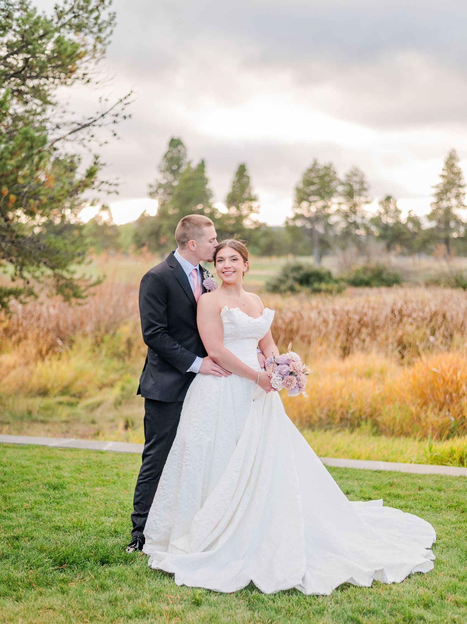 Bride in white gown holding pink bouquet stands with groom in dark suit kissing her cheek at outdoor wedding.