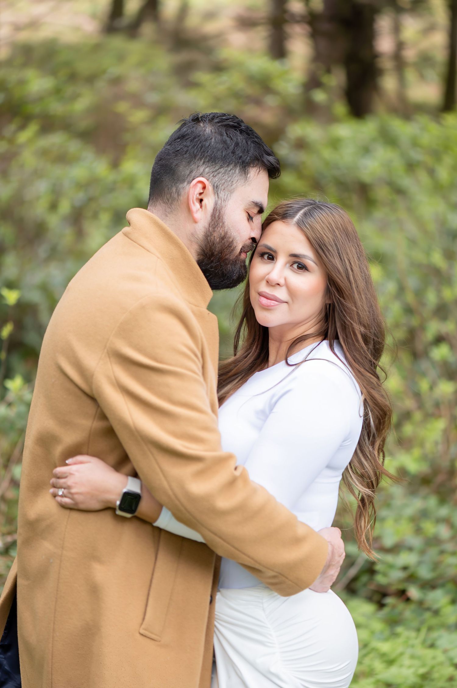 Couple embracing in a lush green forest, woman in white dress smiling at camera while man kisses her cheek.