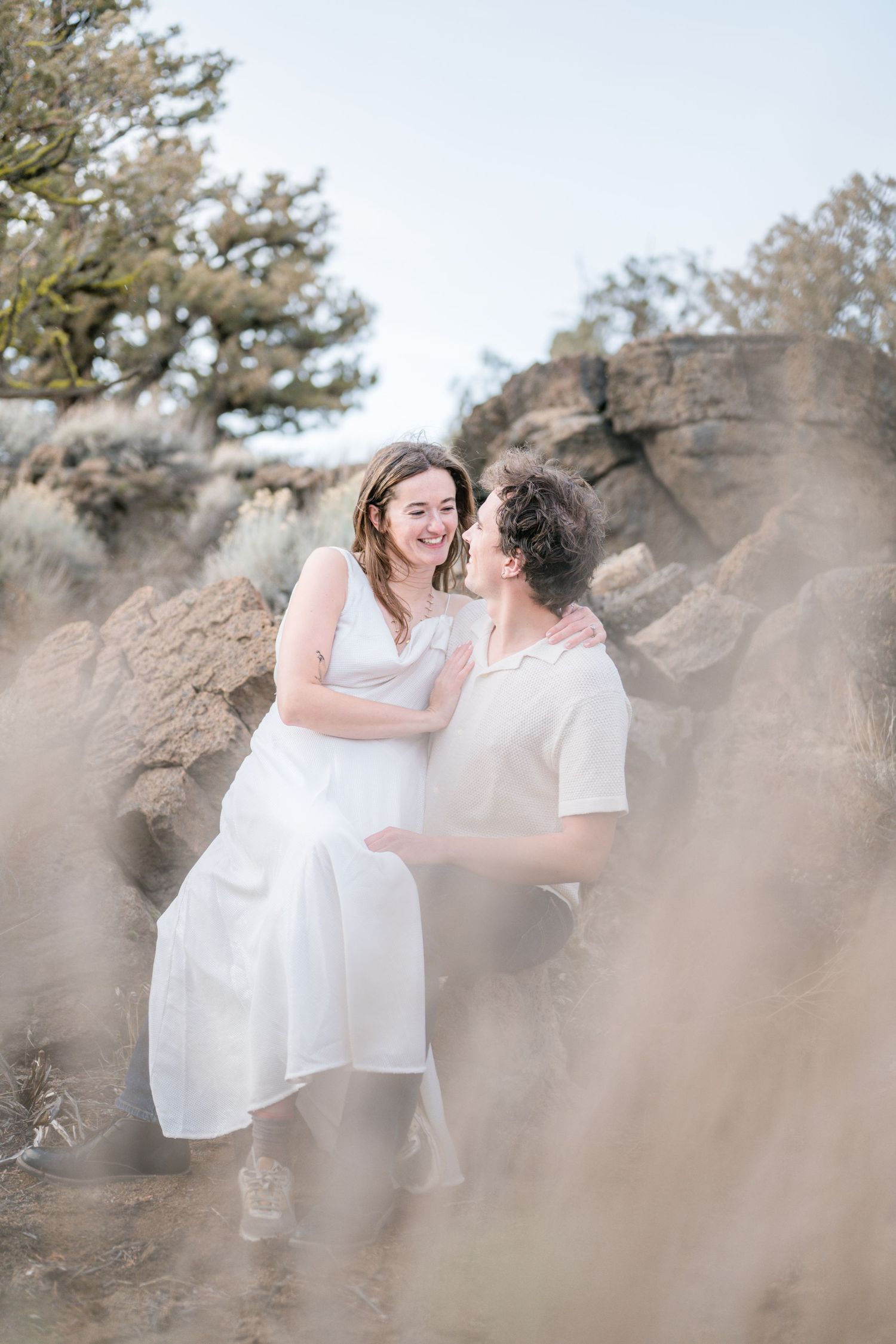 A couple in white outfits embraces and smiles among rocky desert terrain with soft, dreamy lighting.