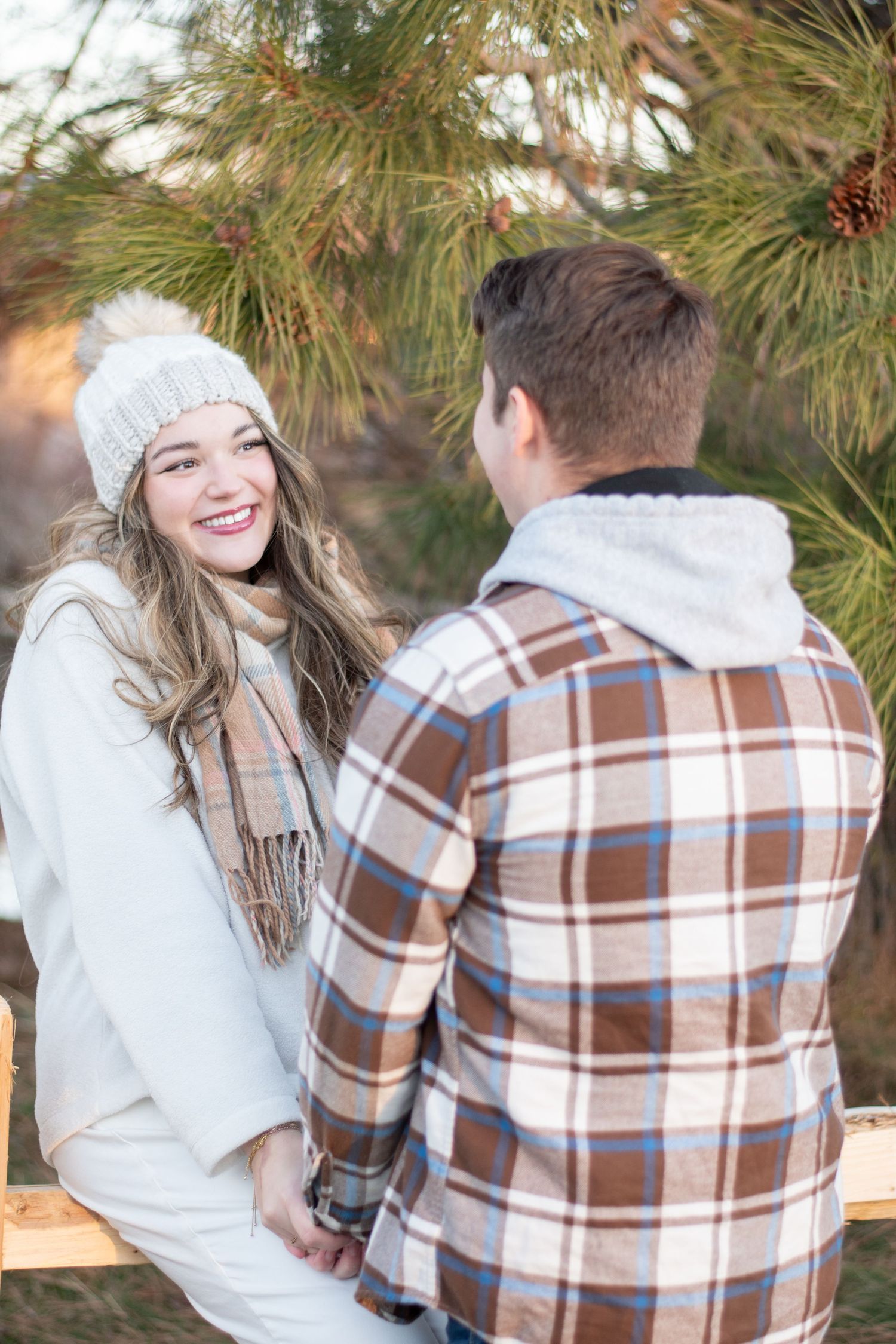 Playful winter engagement photos with couple in plaid and white under pine trees.