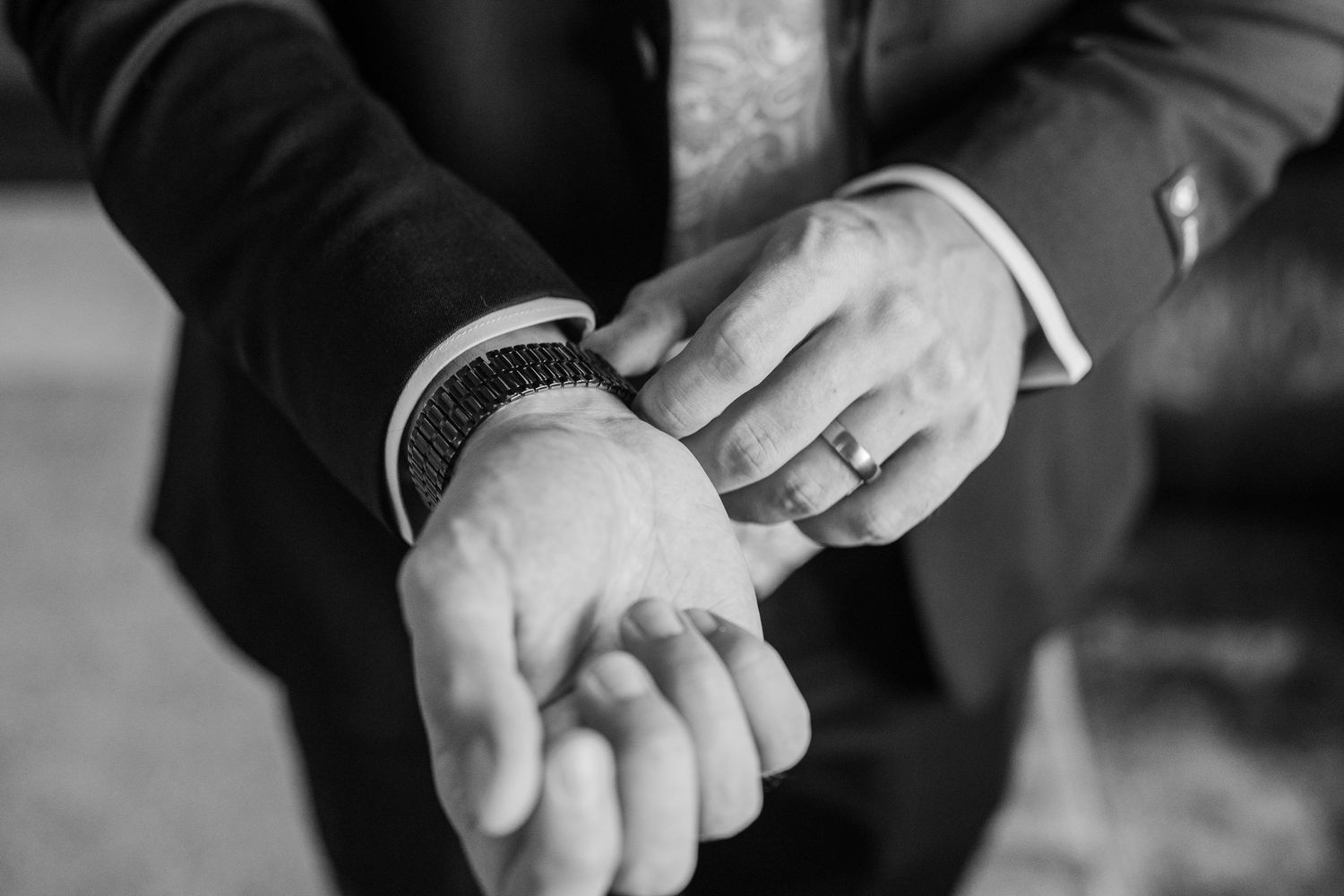 Black and white photo of a groom adjusting his watch cufflinks wearing a wedding ring and dark suit.