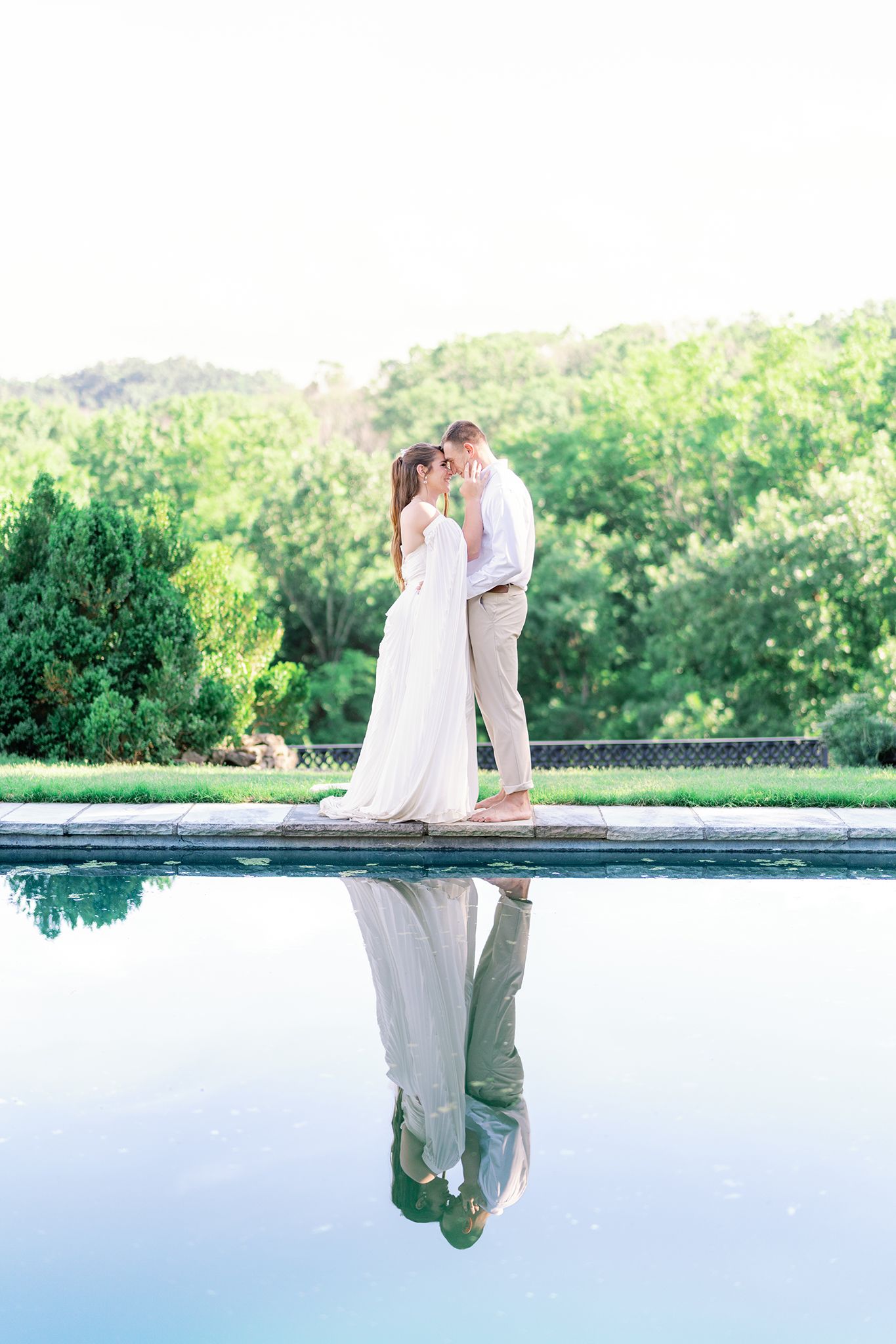 Couple in wedding attire sharing a romantic moment by a reflective pool with lush greenery backdrop.