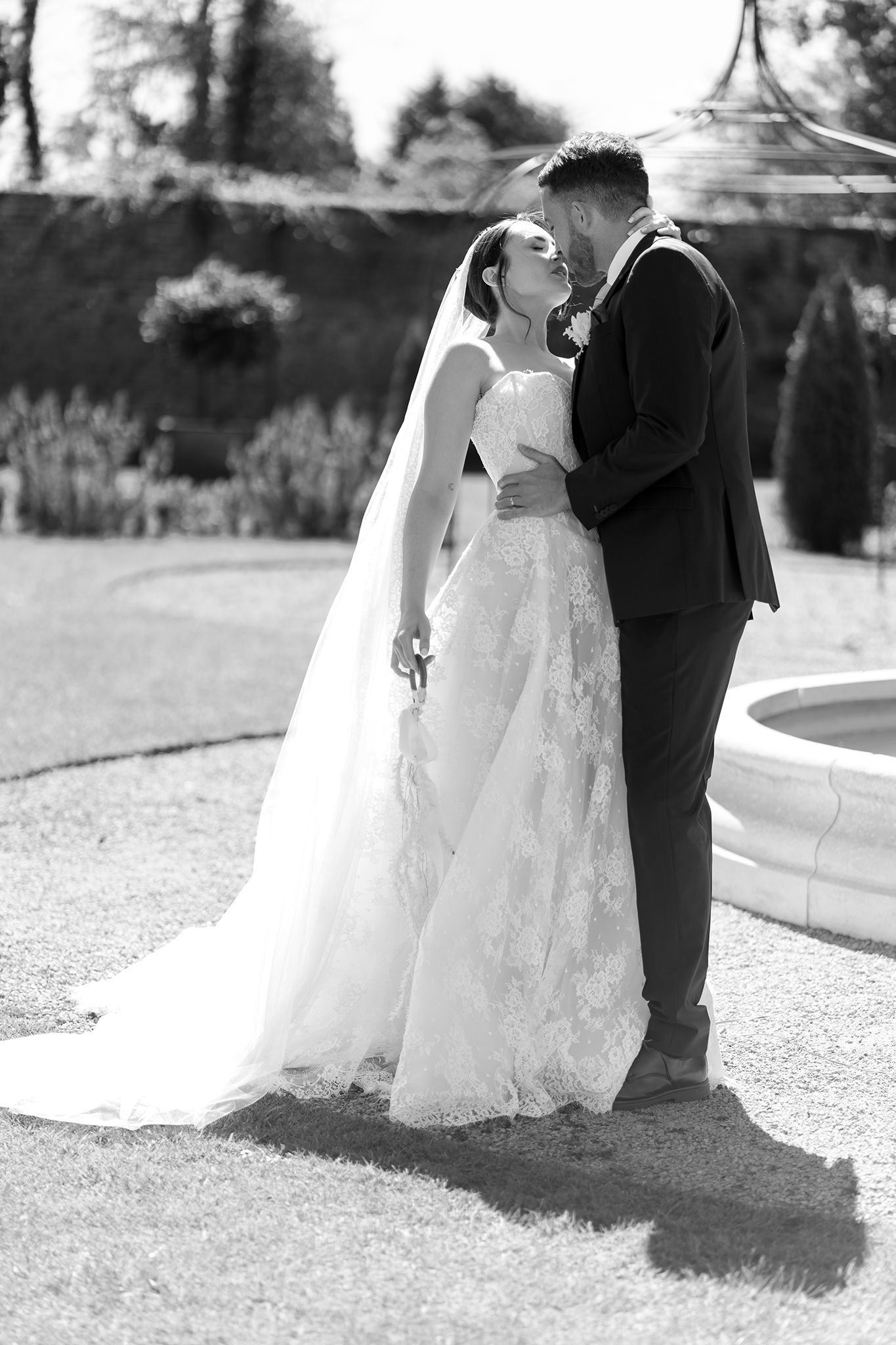 Black and white photo of a bride and groom sharing a romantic moment in a garden, bride wearing a lace gown with veil.