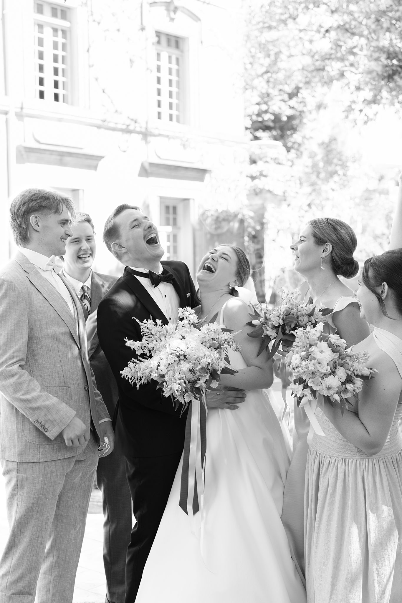 Black and white wedding party photo with bride and groom laughing joyfully alongside bridesmaids and groomsmen outdoors.