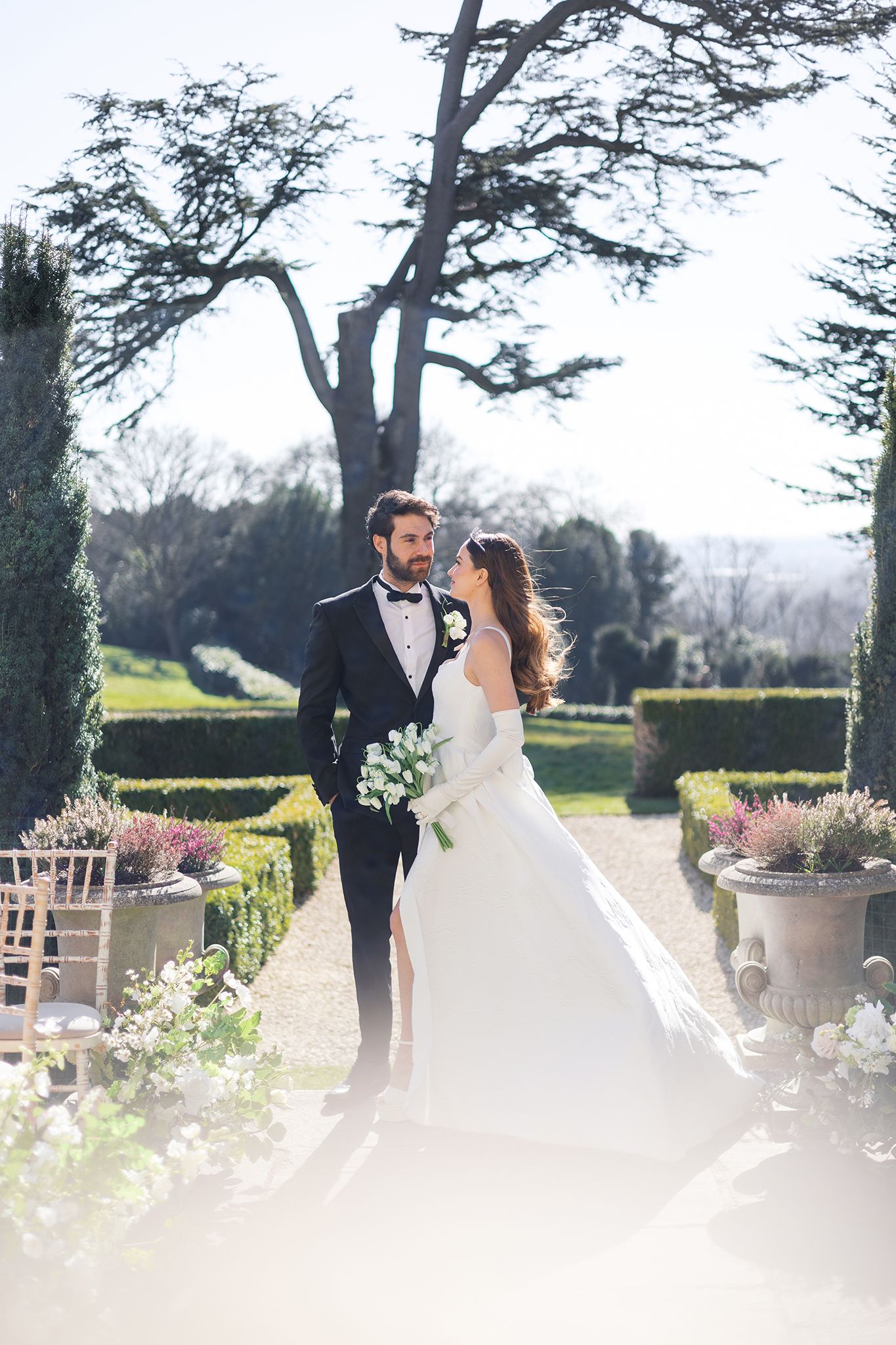 A couple in wedding attire stands on a garden path surrounded by manicured hedges and elegant stone planters.
