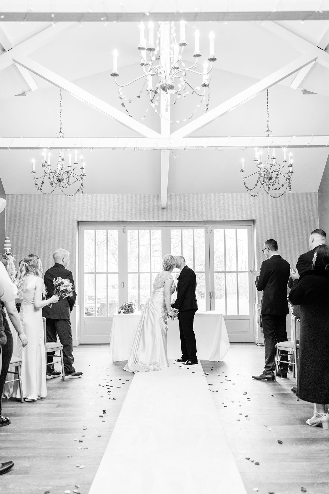 Black and white photo of an elegant wedding ceremony inside a chapel with crystal chandeliers and scattered rose petals.