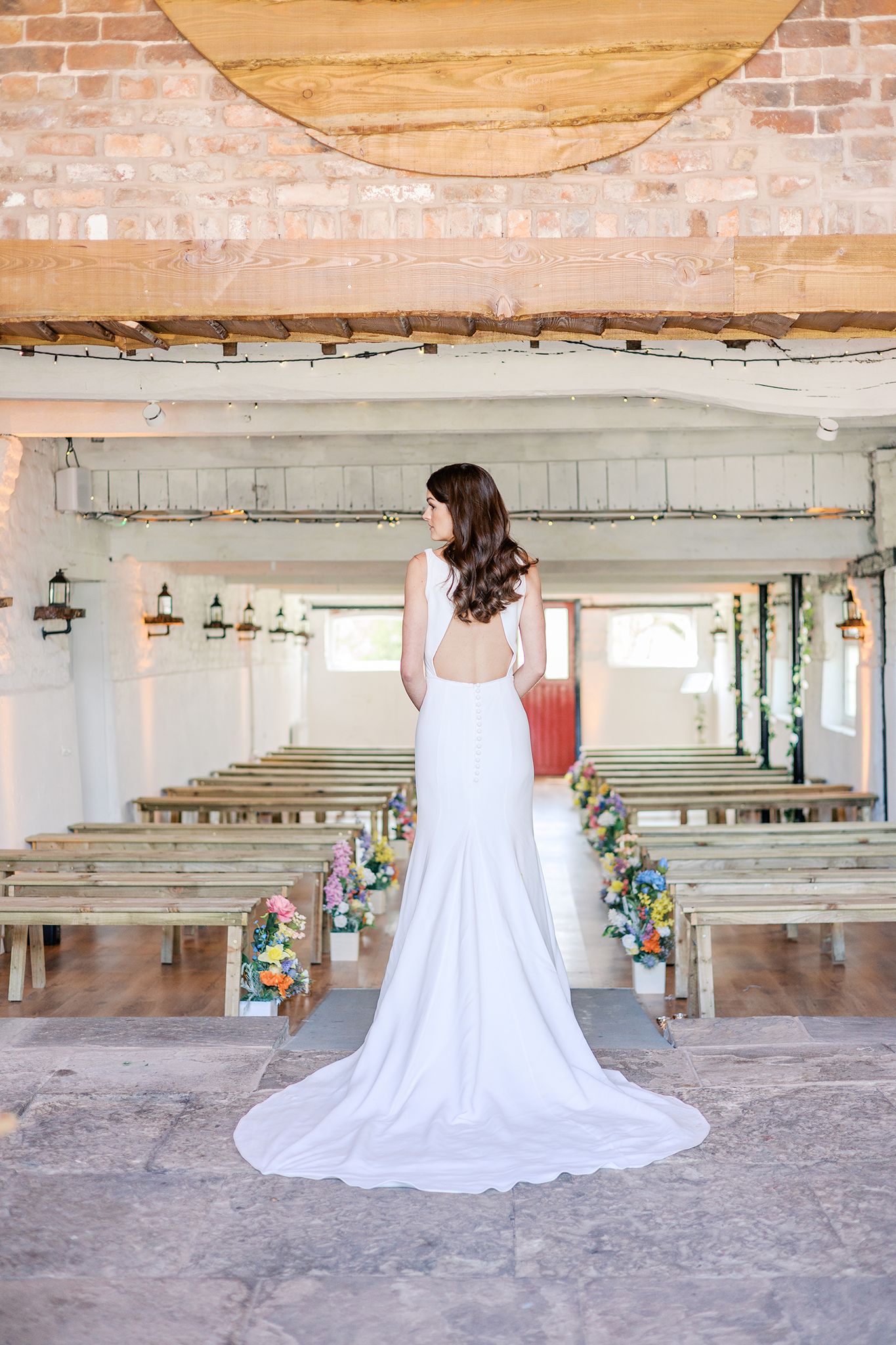 A bride in a flowing white dress stands in a rustic venue with exposed brick walls and wooden benches adorned with flowers.