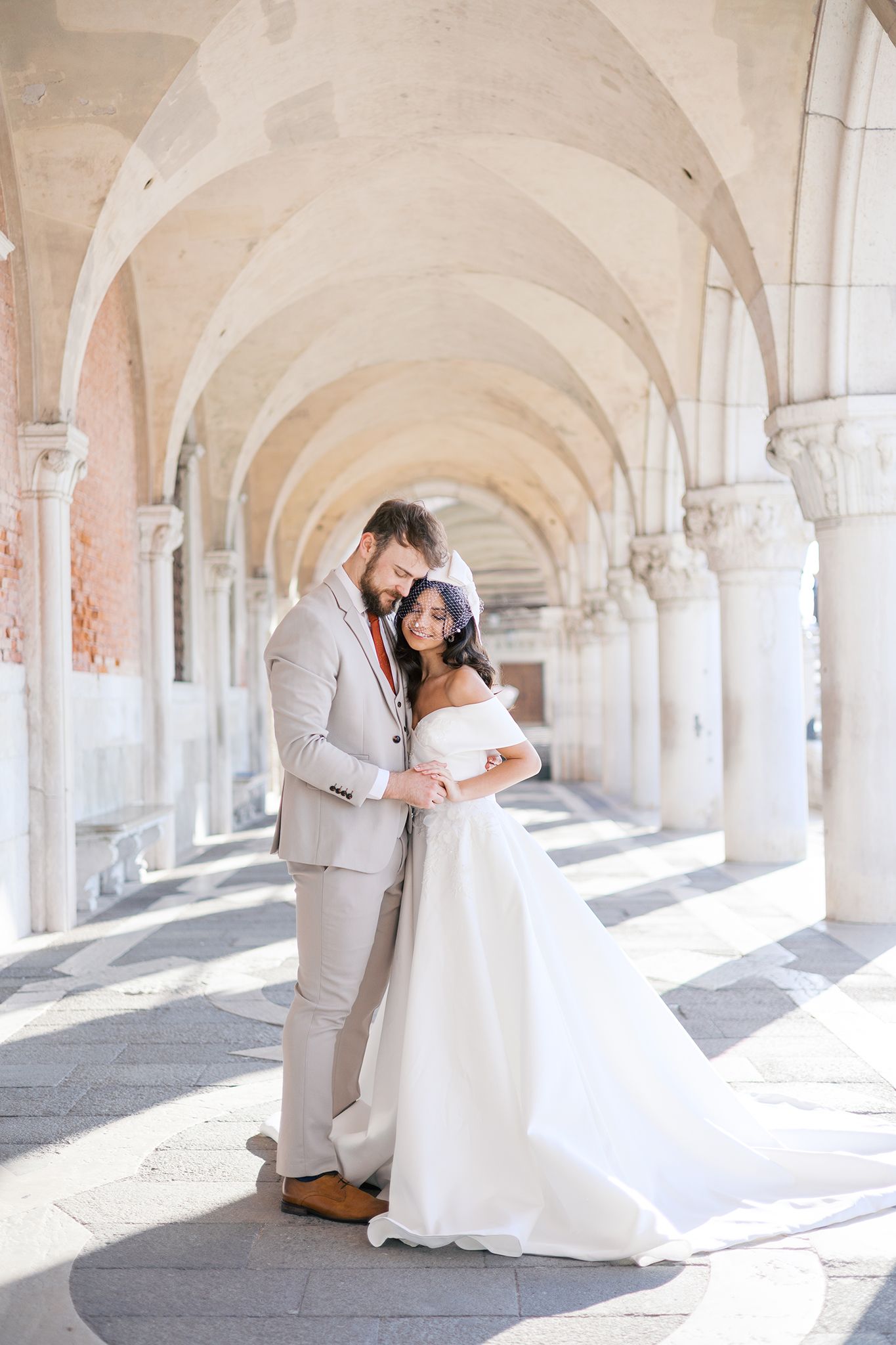 A couple in wedding attire embraces under elegant white stone archways in a bright sunlit corridor.