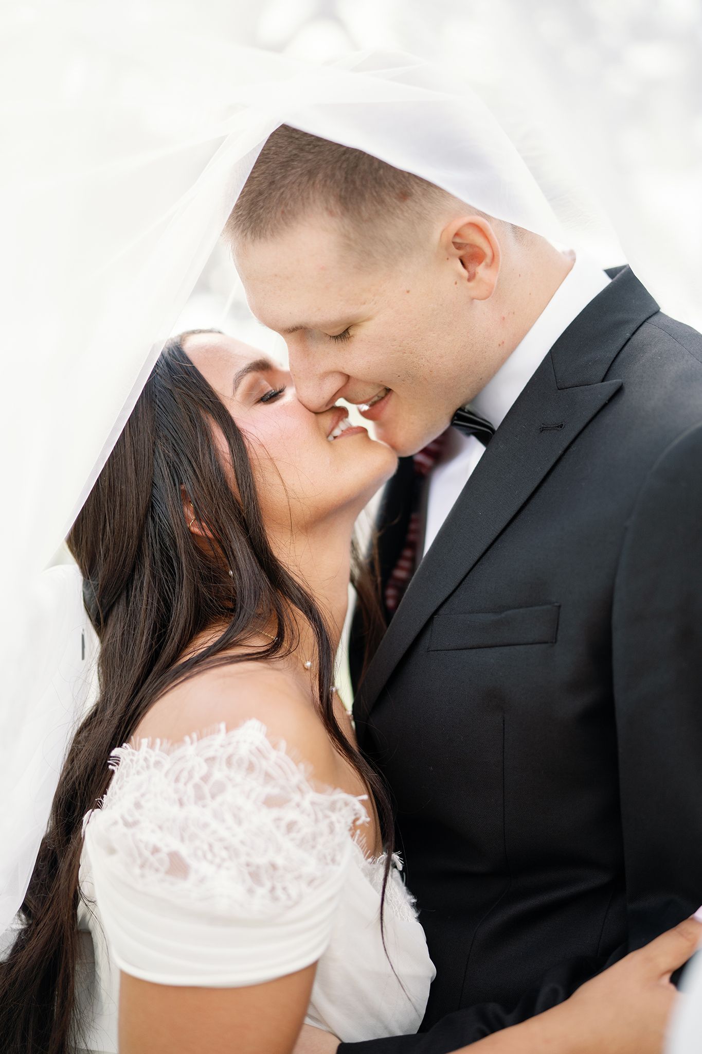 Bride and groom sharing a romantic kiss under a white veil on their wedding day.