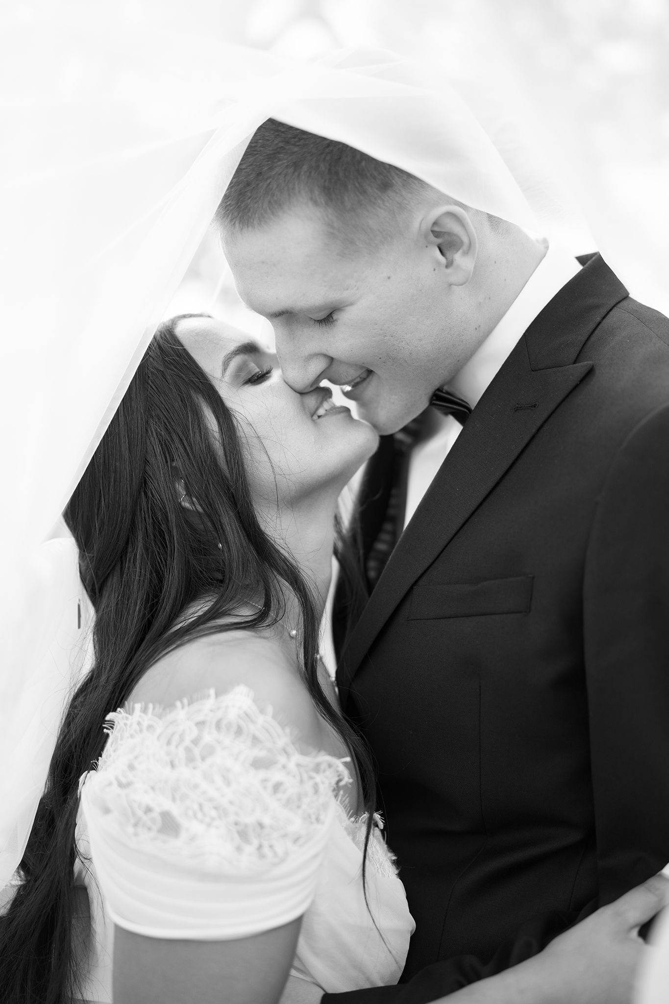 Black and white photo of a bride and groom sharing a kiss under her veil on their wedding day.