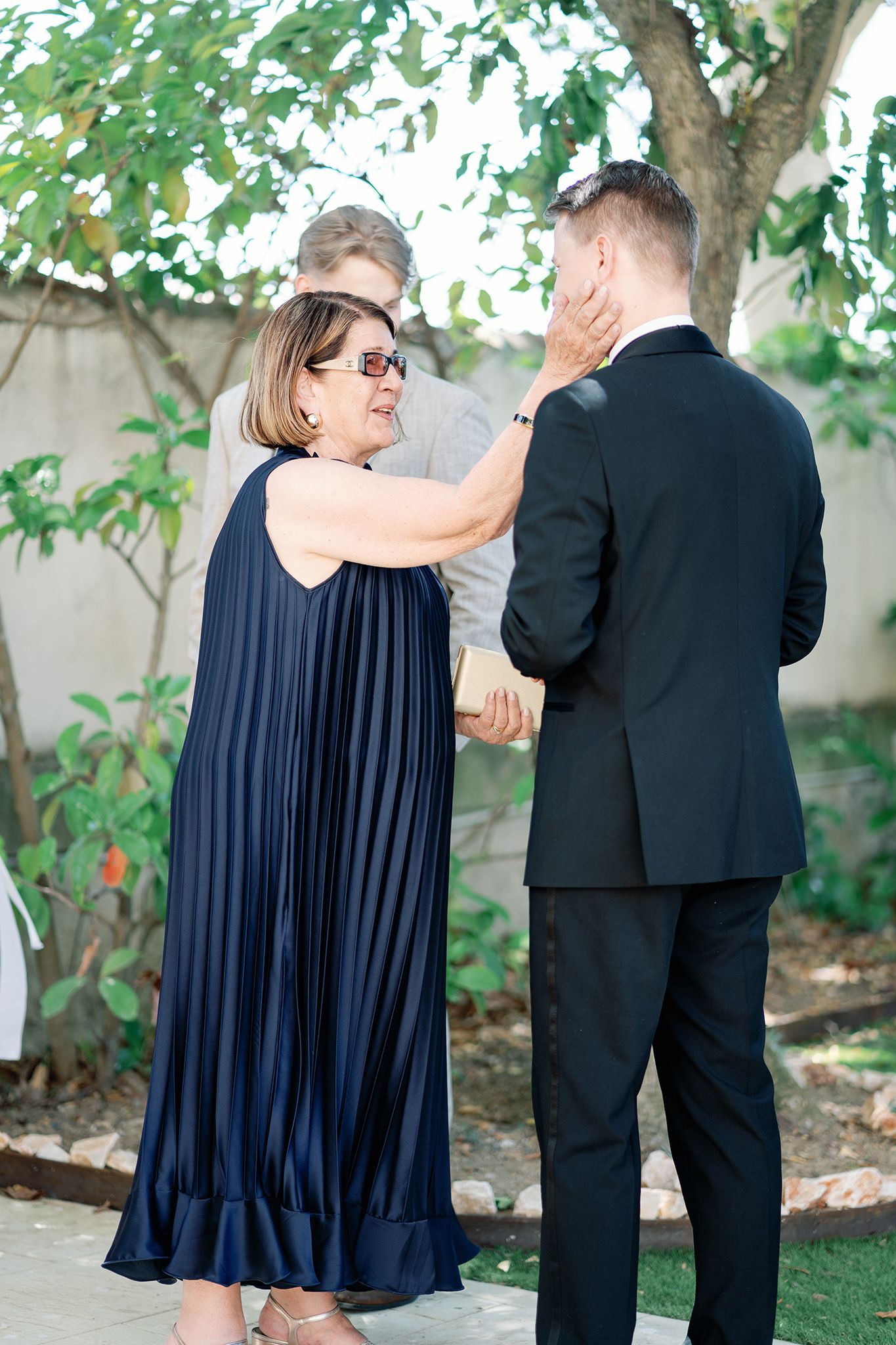 A woman in a navy pleated dress touches a groom's face outdoors at a wedding ceremony.