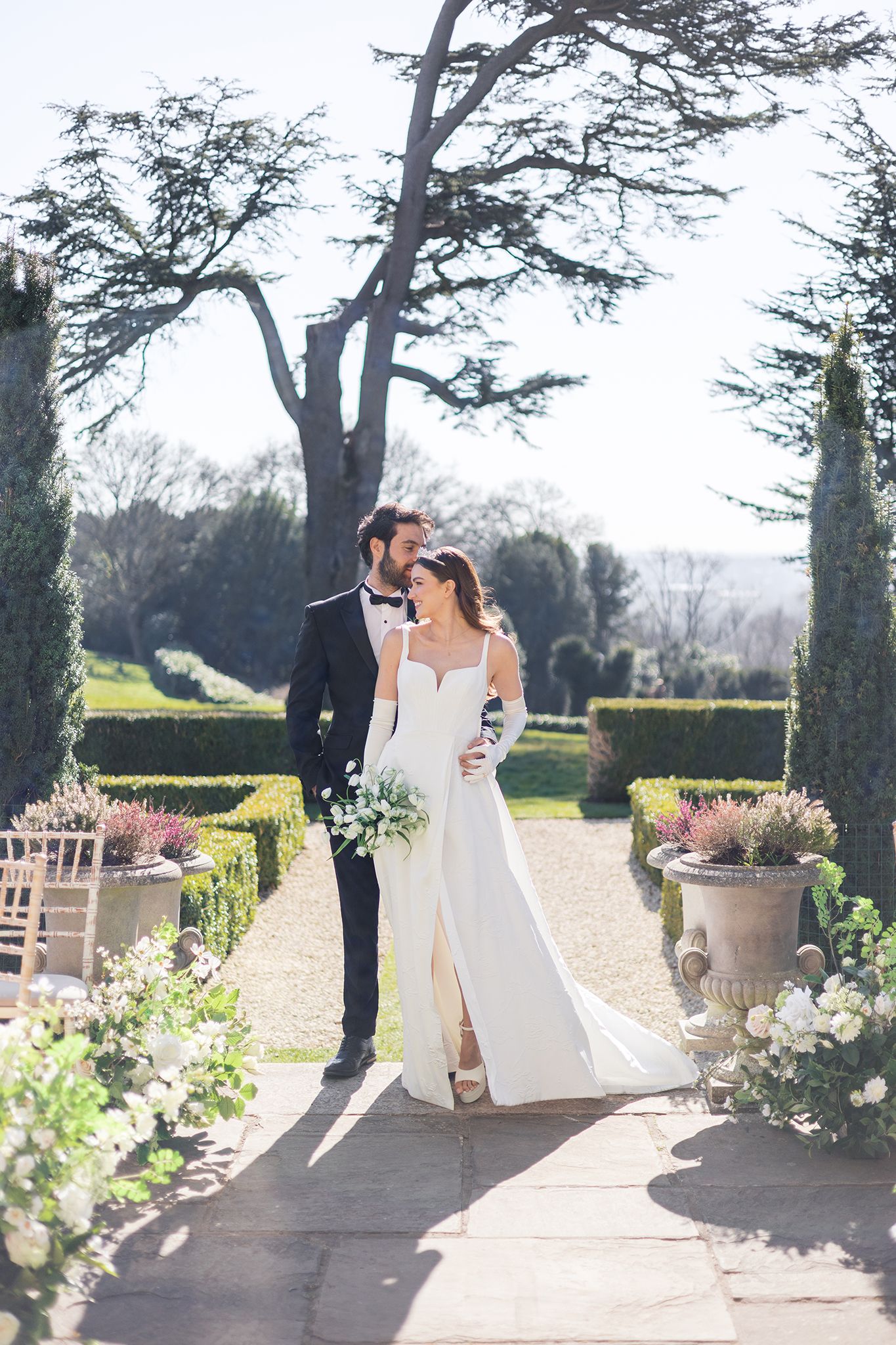 A couple embraces in a romantic garden setting surrounded by manicured hedges and tall trees on a sunny spring day.