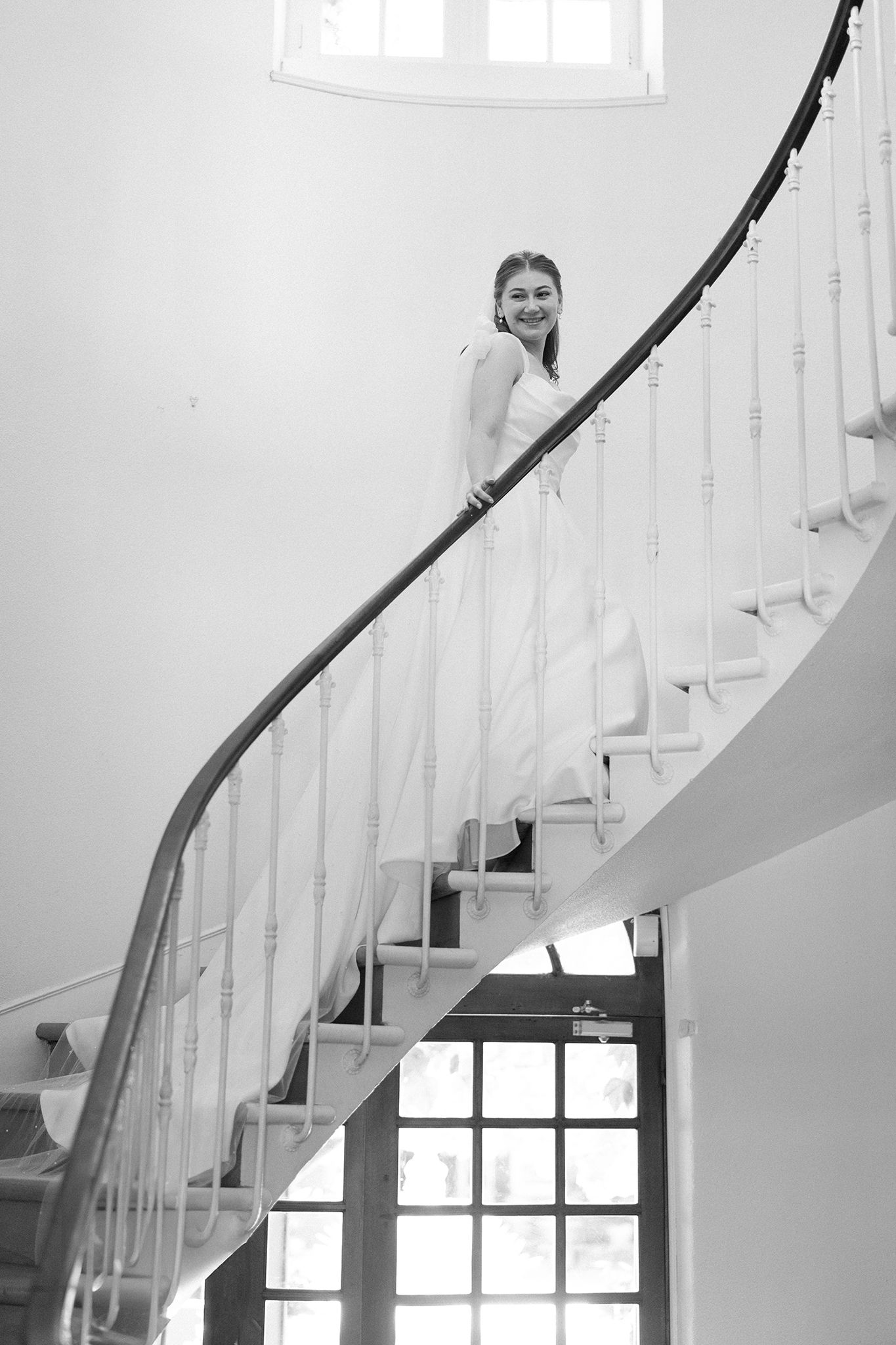 Black and white photo of a smiling woman in a white dress posing on an elegant curved staircase with white railings.