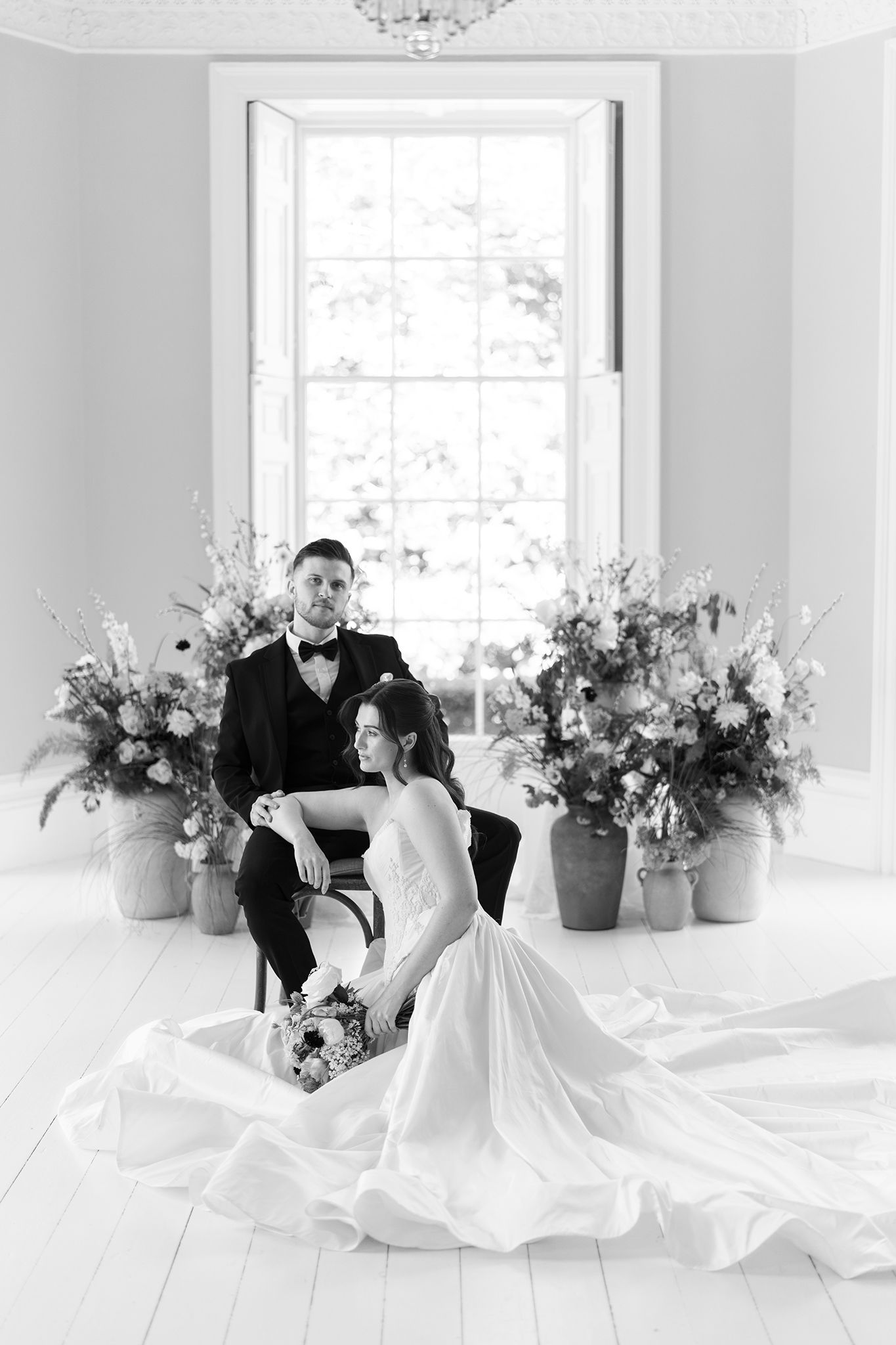 Black and white wedding photo of bride sitting on floor in gown beside groom in tuxedo, with floral arrangements by bright window.