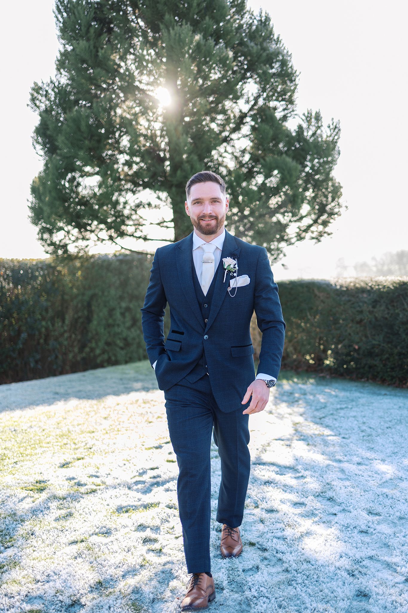 A groom in a navy blue suit walks on a frosty ground with sunlight peeking through a pine tree behind him.