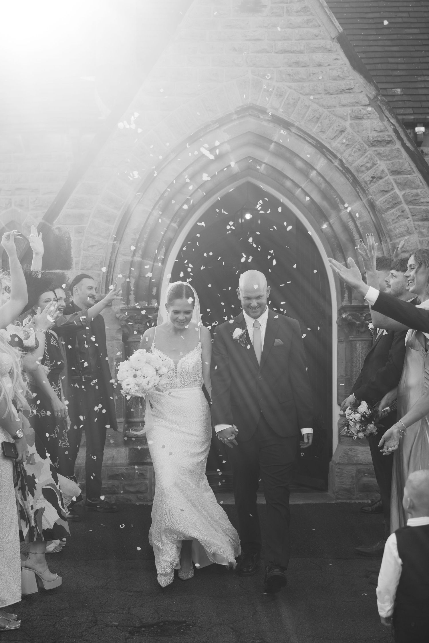 Black and white photo sequence shows wedding couple walking through confetti with guests outside historic brick church archway.