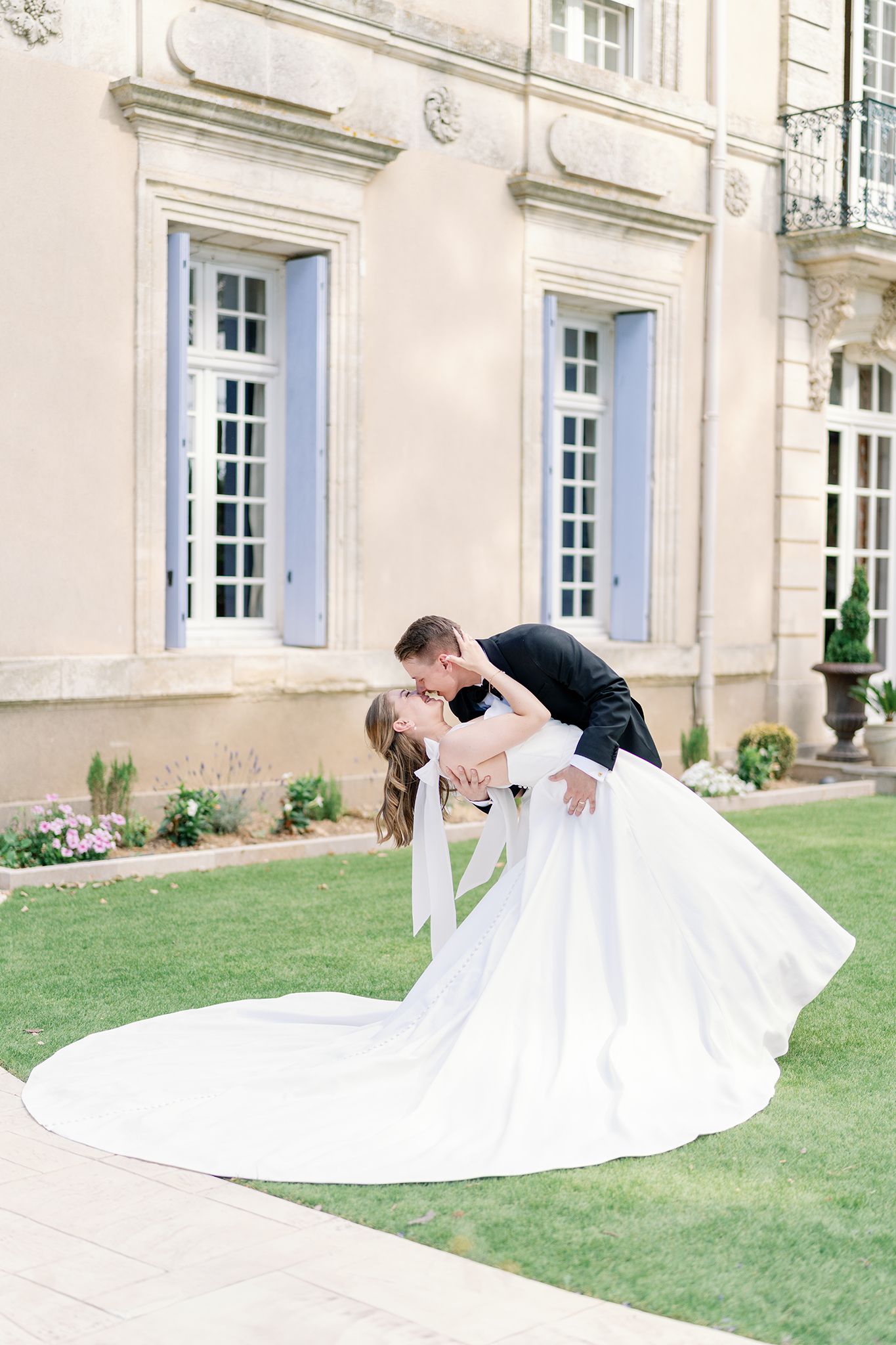 Groom dips bride in elegant white gown for a romantic kiss outside a French chateau on their wedding day.