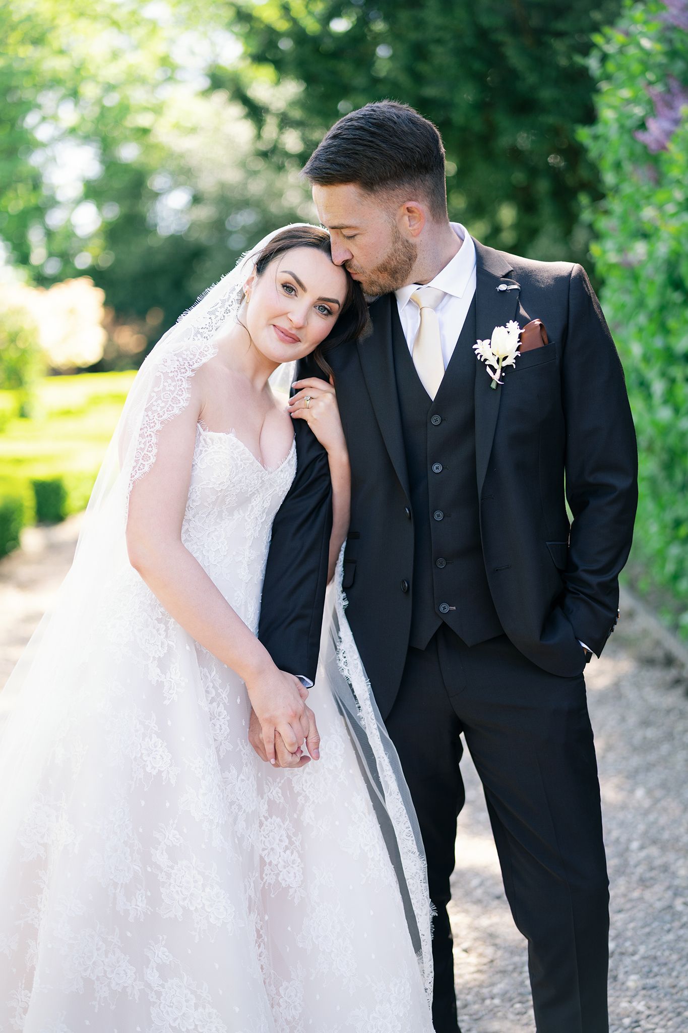 Bride in lace gown and veil leans on groom in black suit as he kisses her head in a sunlit garden.