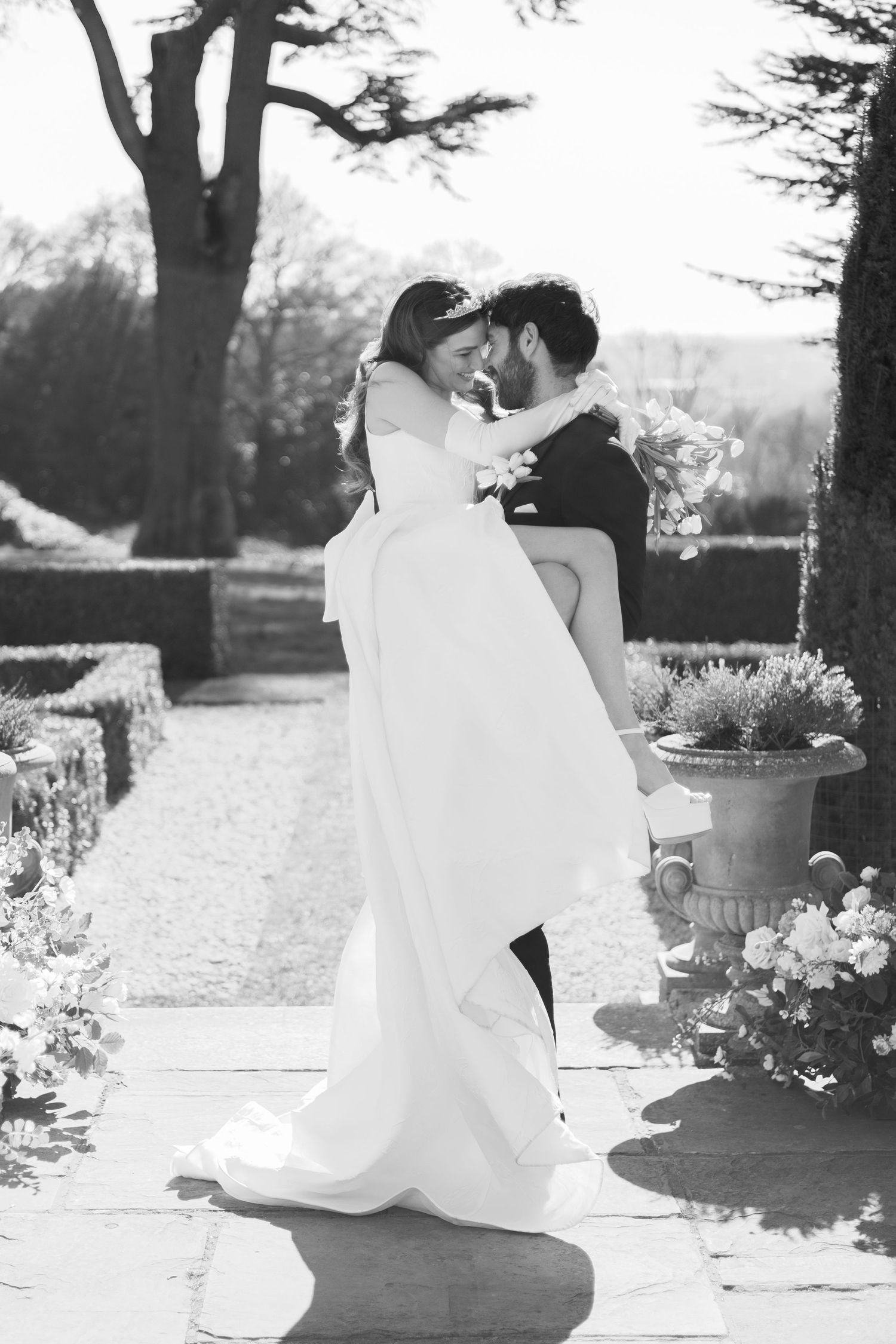 A romantic black and white wedding photograph showing a couple embracing on a garden path surrounded by trees and flowers.