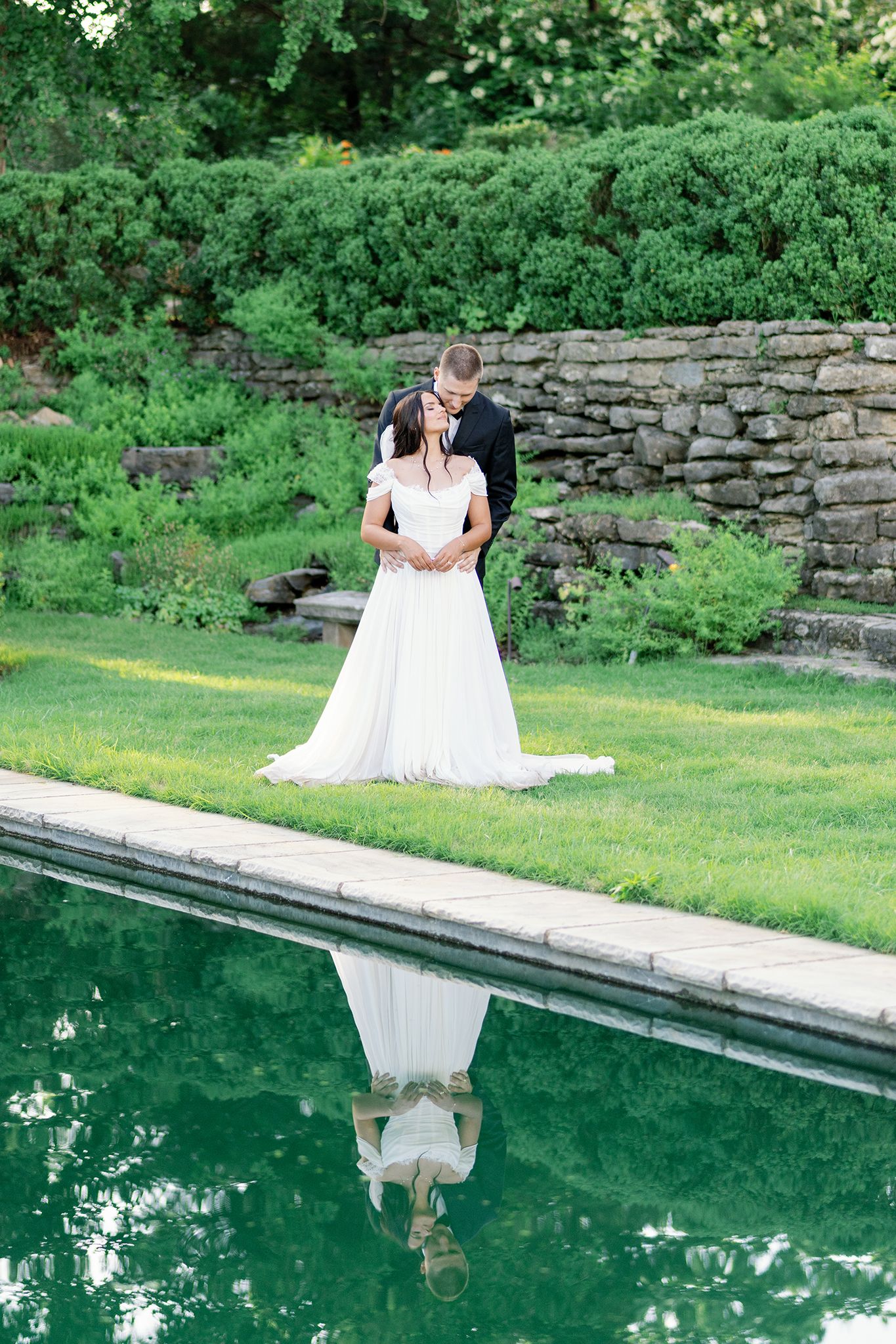 Bride and groom embrace by a reflective pool in a lush garden, with the couple mirrored in the still water below.