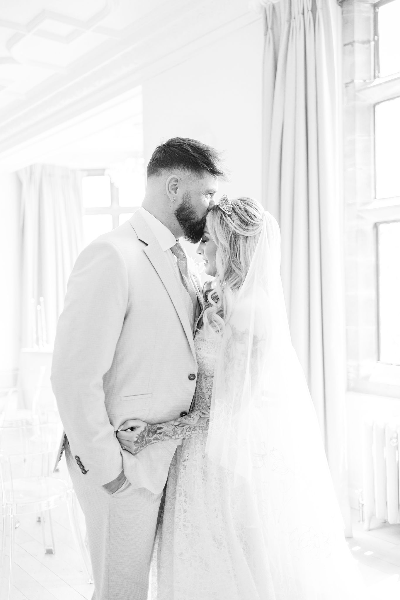 A romantic black and white wedding photo of a couple sharing an intimate moment by a window with light curtains.