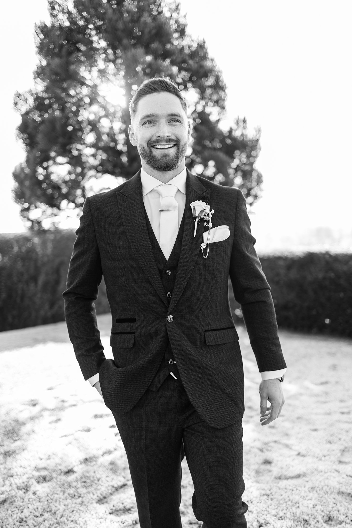 Black and white photo of a smiling groom in a three-piece suit with white tie and boutonniere outdoors.