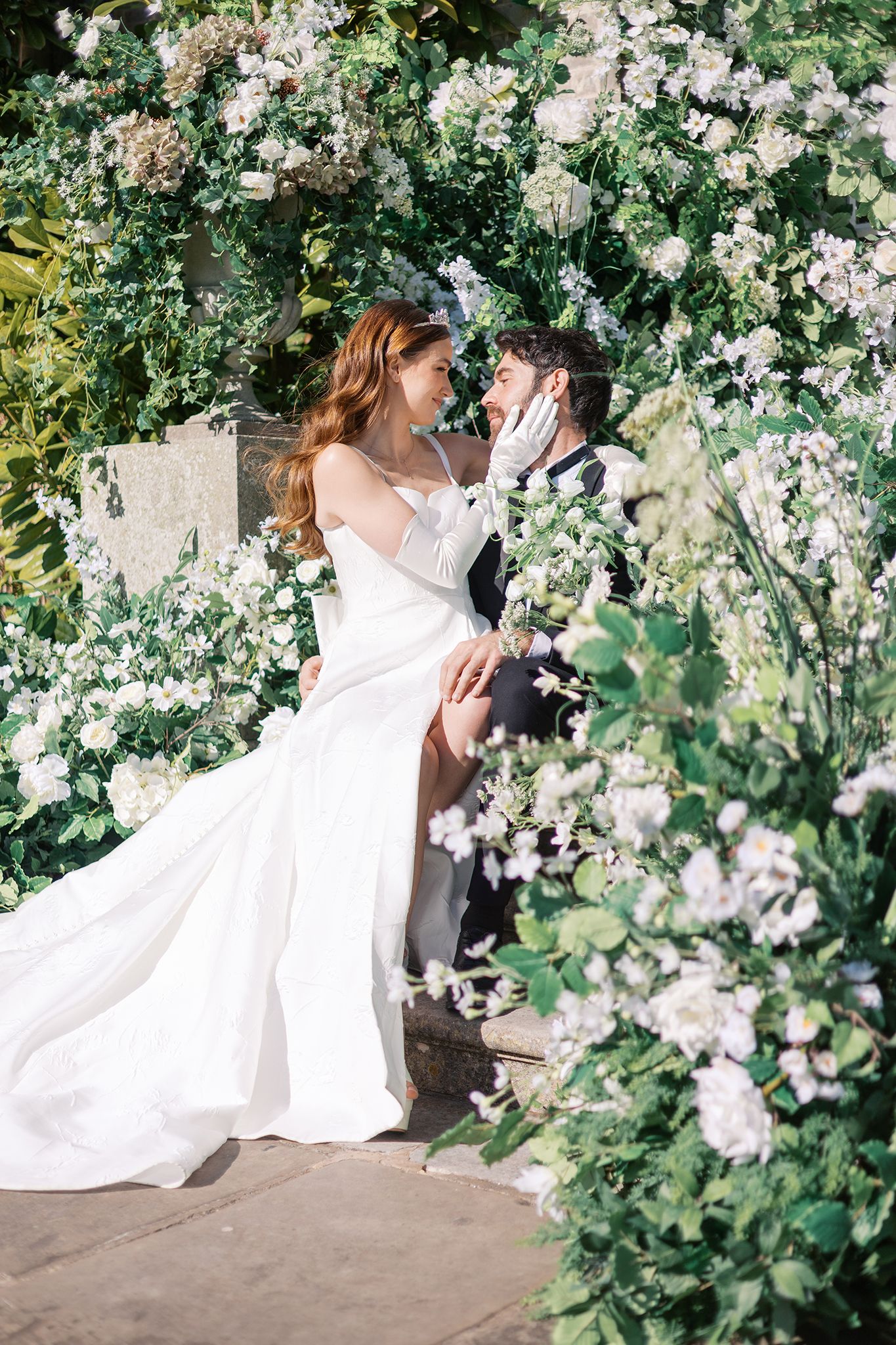 A bride in a flowing white dress poses among blooming white roses in a romantic garden setting.