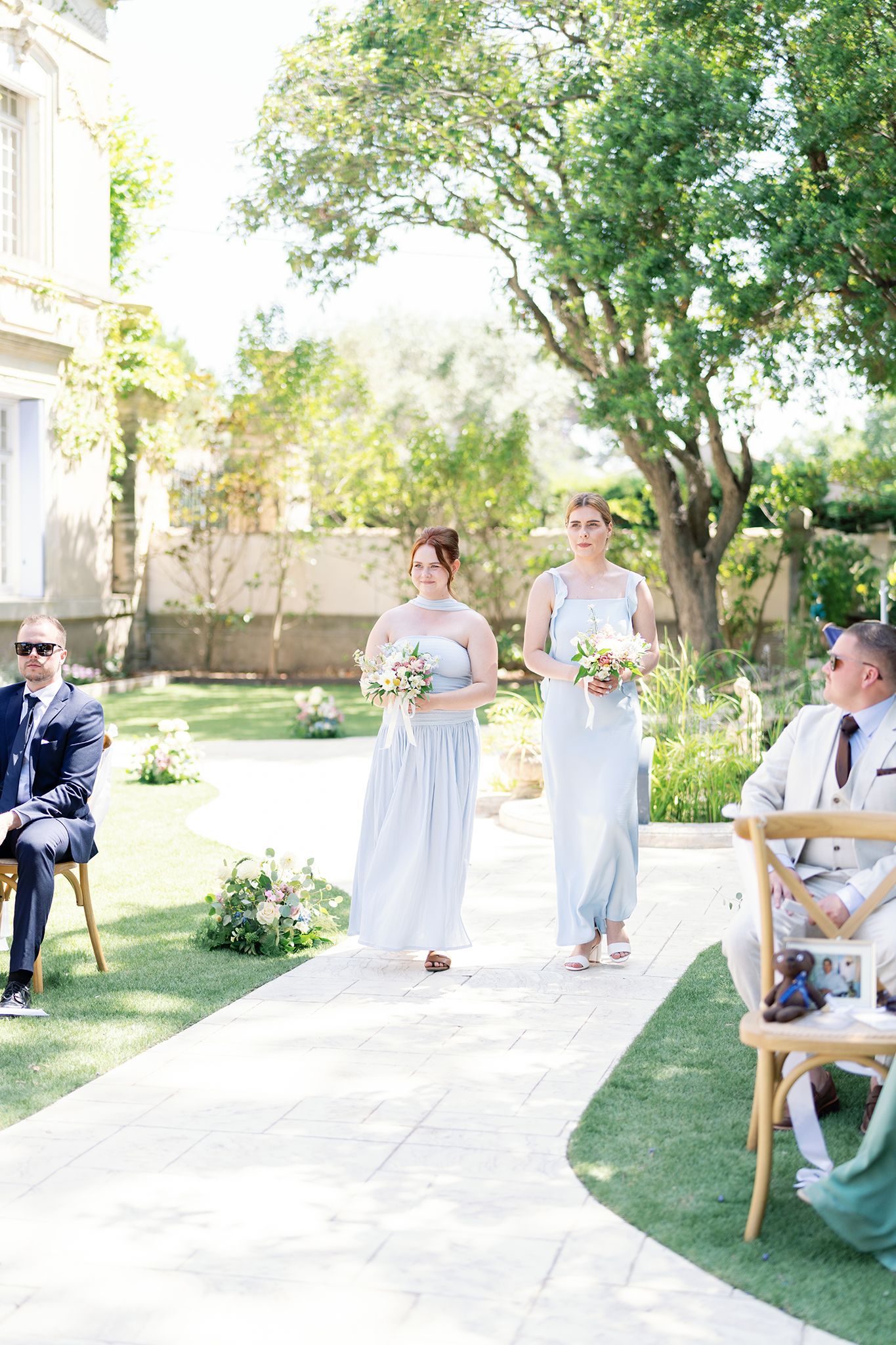 Two bridesmaids in light blue gowns carrying bouquets walk down outdoor wedding aisle surrounded by lush garden greenery.