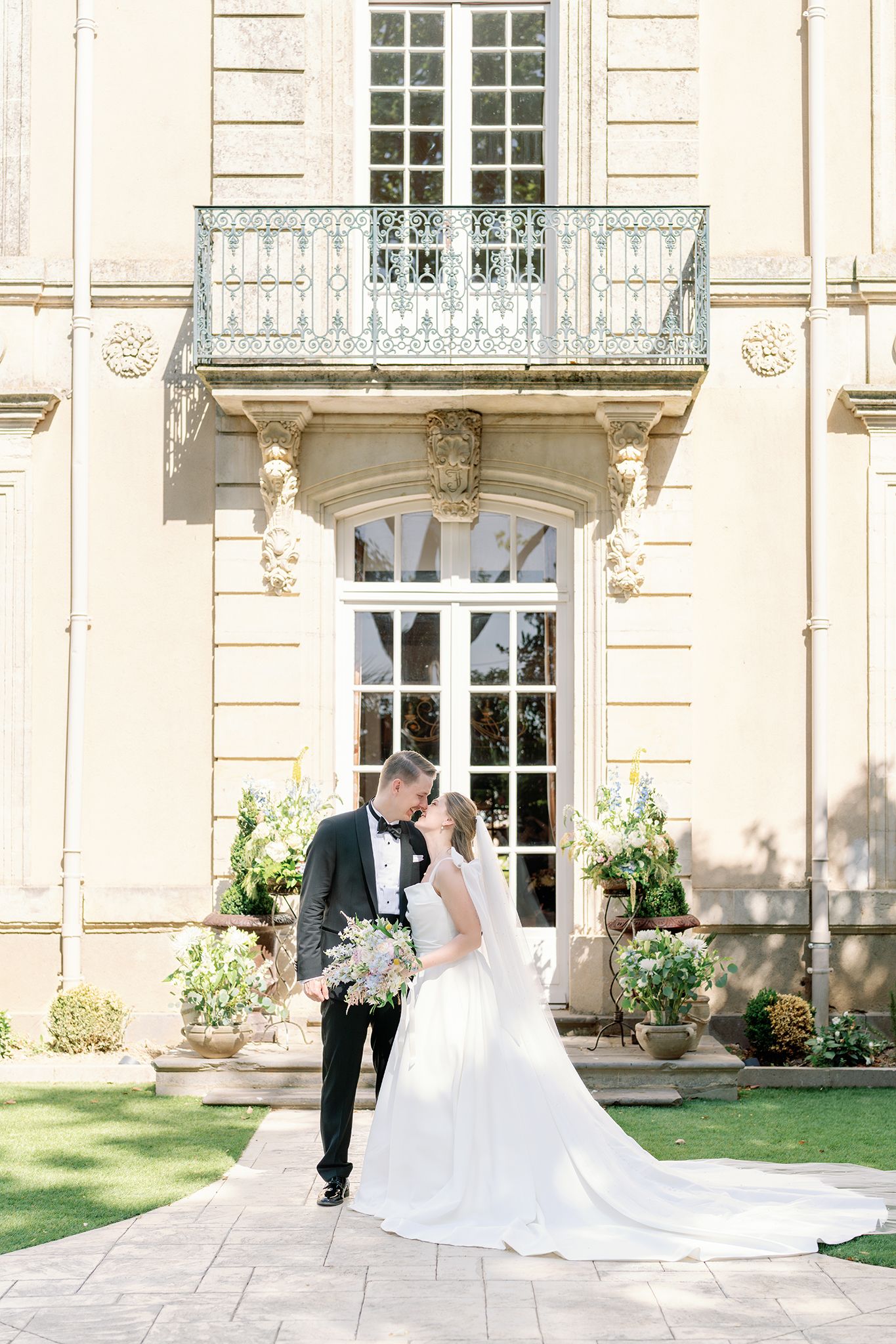 Bride and groom share a romantic kiss outside an elegant French château with ornate balcony and lush garden florals.