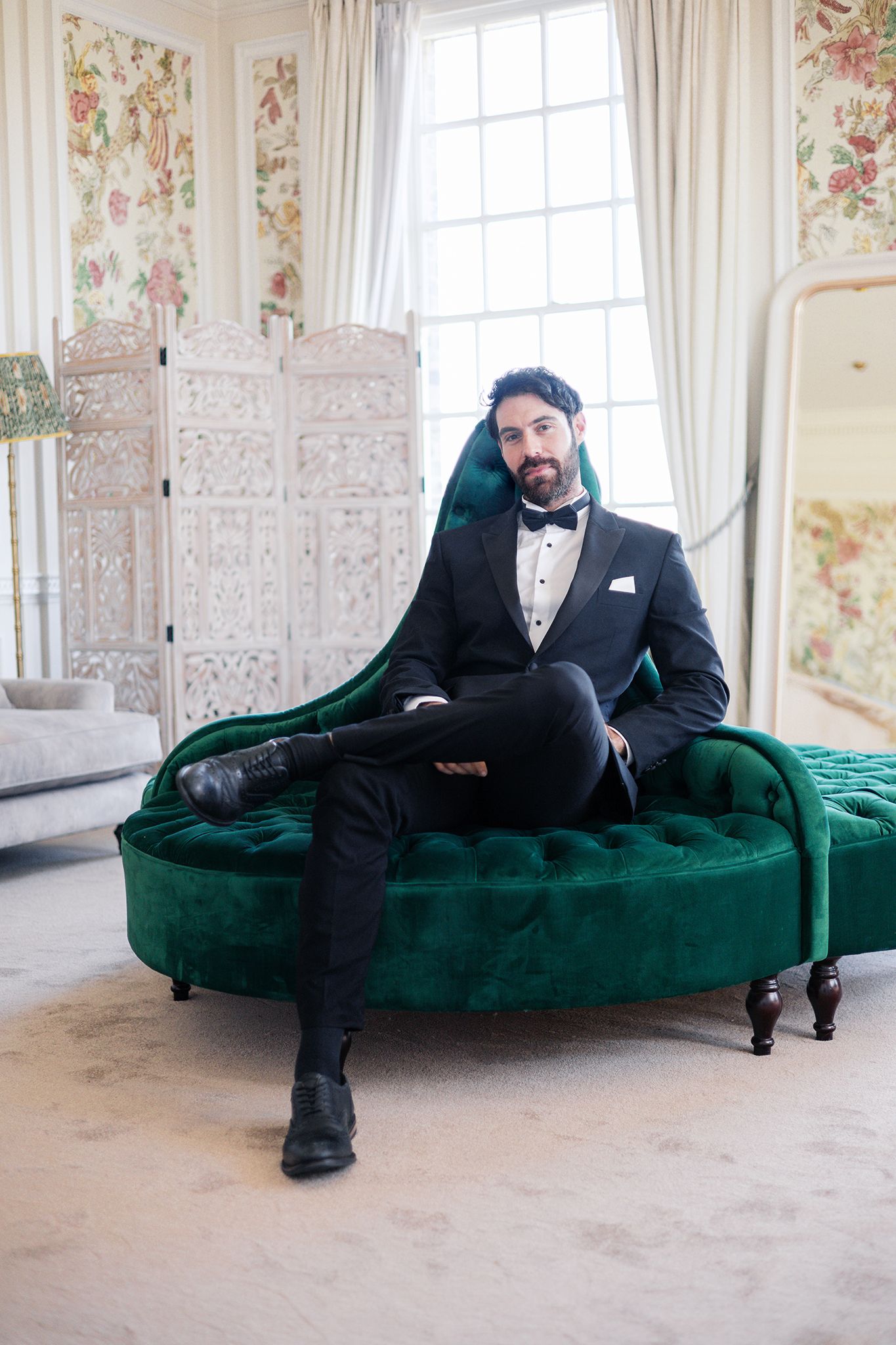 Elegant formal attire worn by someone lounging on a luxurious emerald green velvet chaise in a bright room with floral wallpaper.