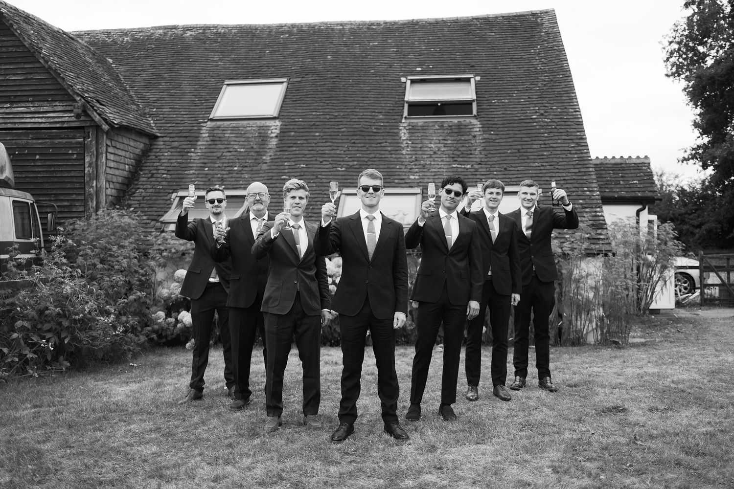 Wedding party members in formal black attire pose in a line outdoors against rustic farm buildings.