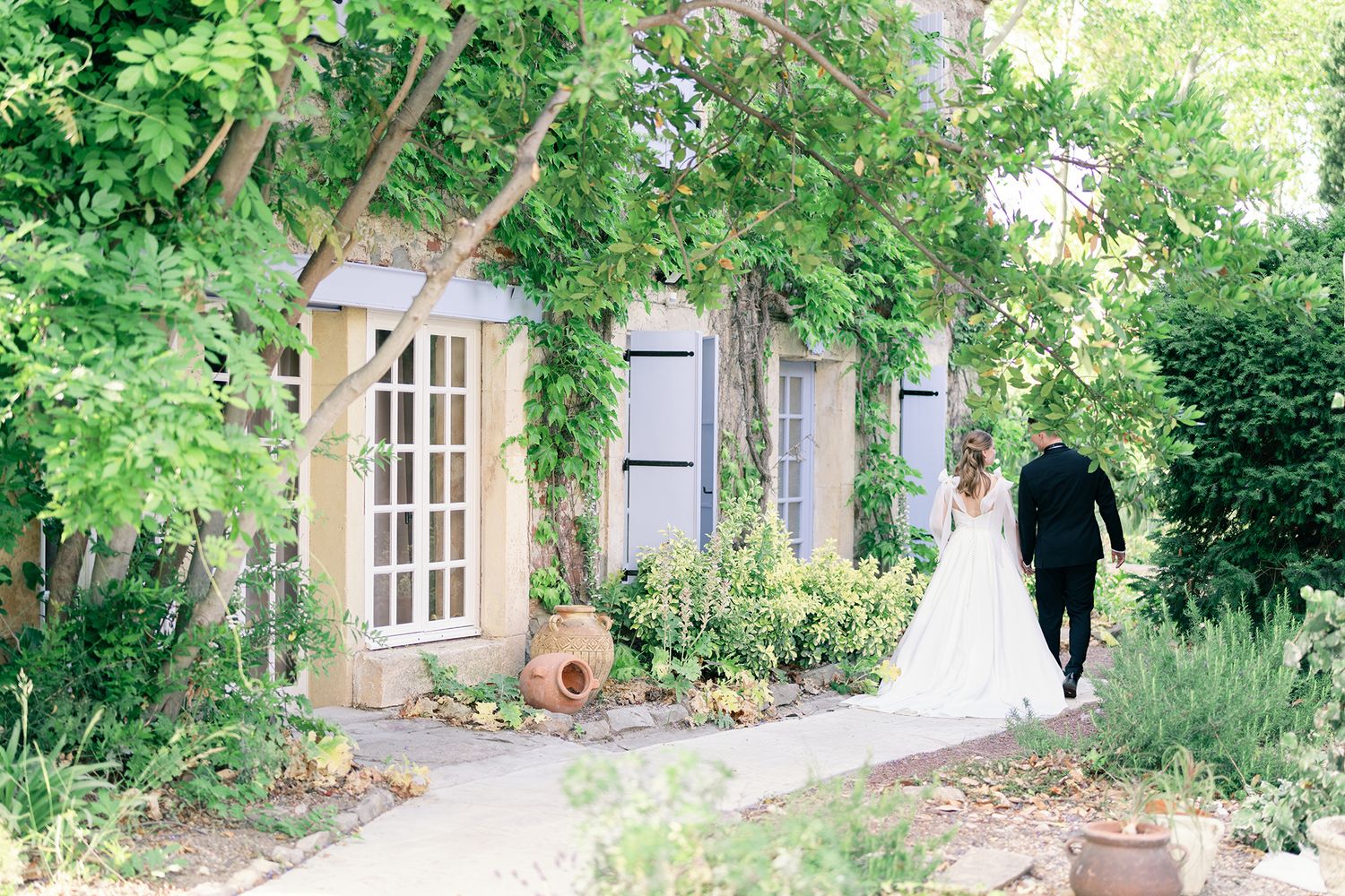 Bride and groom walking hand in hand toward a charming stone cottage surrounded by lush greenery on their wedding day.