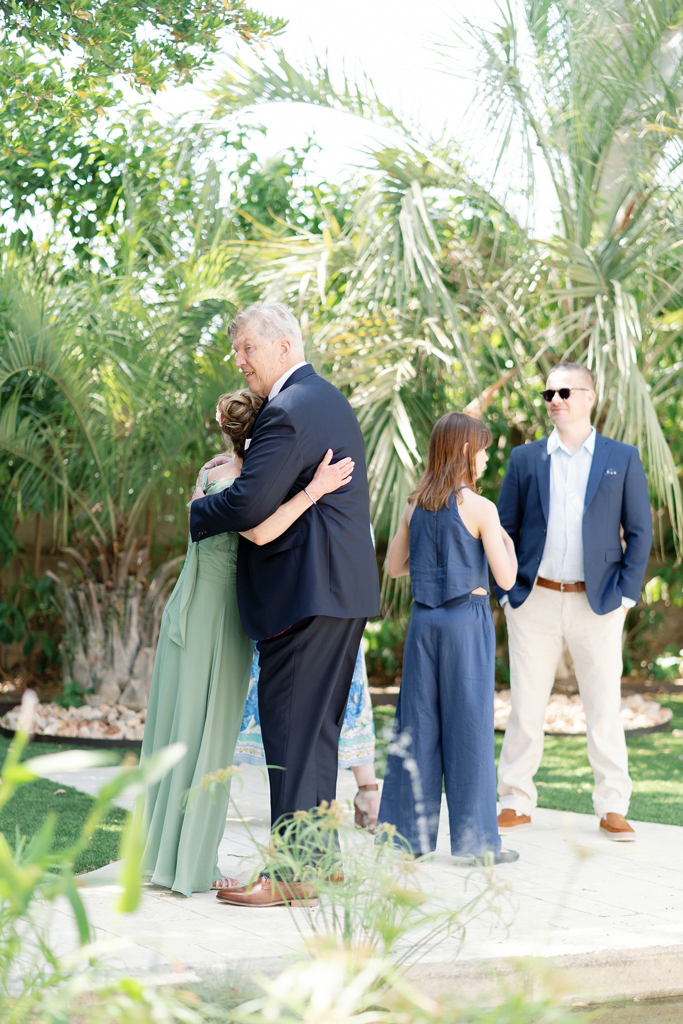Guests hugging and socializing outdoors at a garden wedding reception surrounded by lush tropical palm trees.
