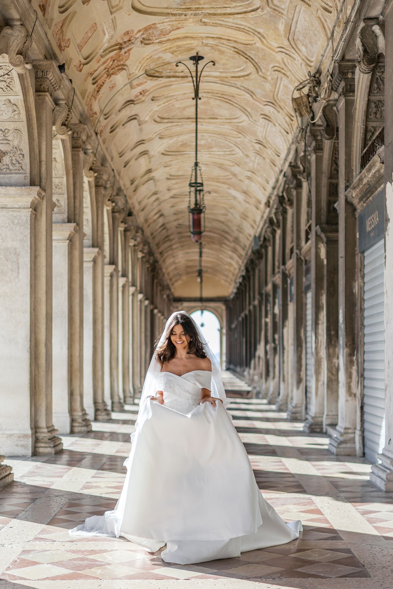 A bride in an off-shoulder white wedding gown poses in an elegant arched colonnade with ornate ceiling and marble floors.
